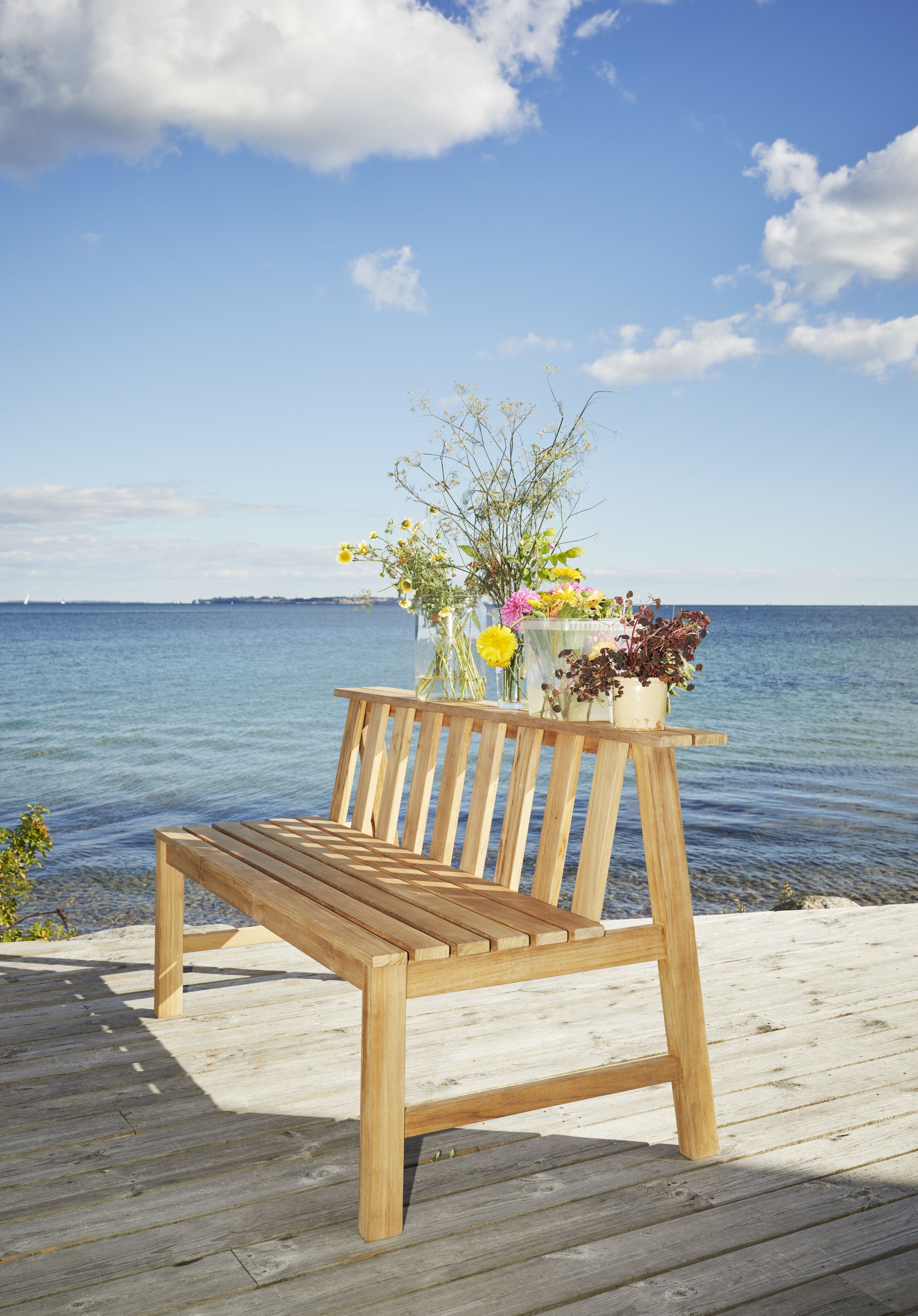 Cadence Teak Bank von Skagerak by Fritz Hansen auf Holzterrasse mit Meerblick und Blumen.