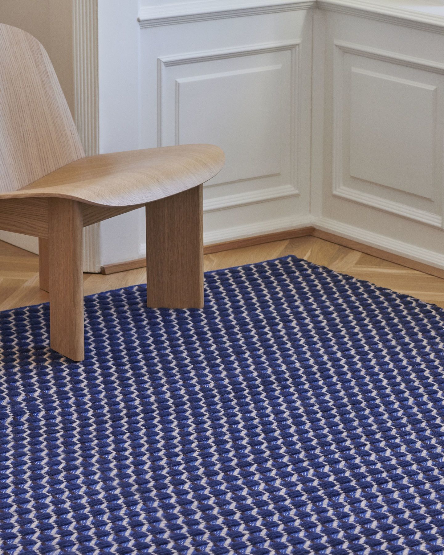 Nahaufnahme des blau-weißen Channel Rug Teppichs von Hay, kombiniert mit einem hellen Holzstuhl auf Parkettboden.