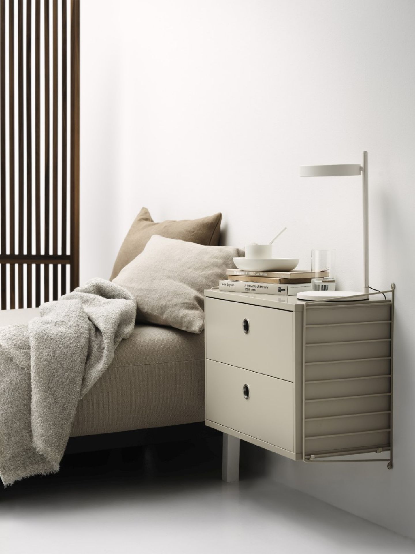 Beige Nachttisch mit zwei Schubladen, Lampe, Buch und Dekoration neben einem Bett mit Kissen und Decke.