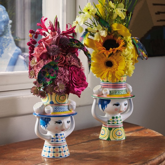 Zwei dekorative Bjorn Wiinblad "Lady with Hat" Vasen mit bunten Blumenarrangements auf einem Holztisch.