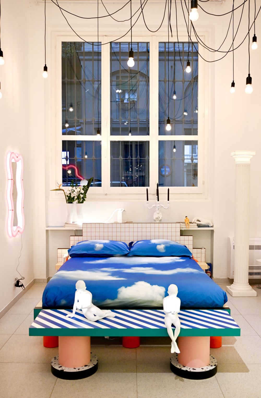 Seletti Bett mit blauem Himmel-Motiv, gestreiftem Bettrahmen und zwei weißen Figuren im Schlafzimmer.