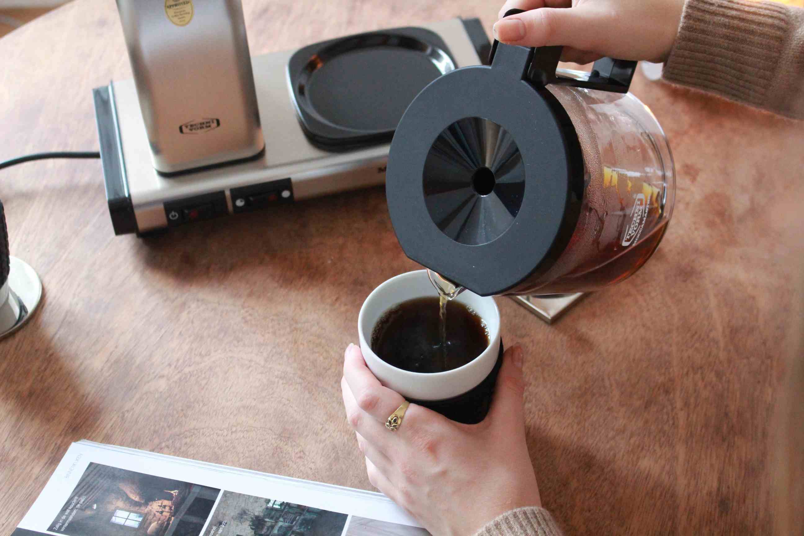 Frau gießt Kaffee aus einer Moccamaster Kaffeemaschine in eine weiße Tasse. Hochwertige Filterkaffeemaschine für Kaffeegenuss.