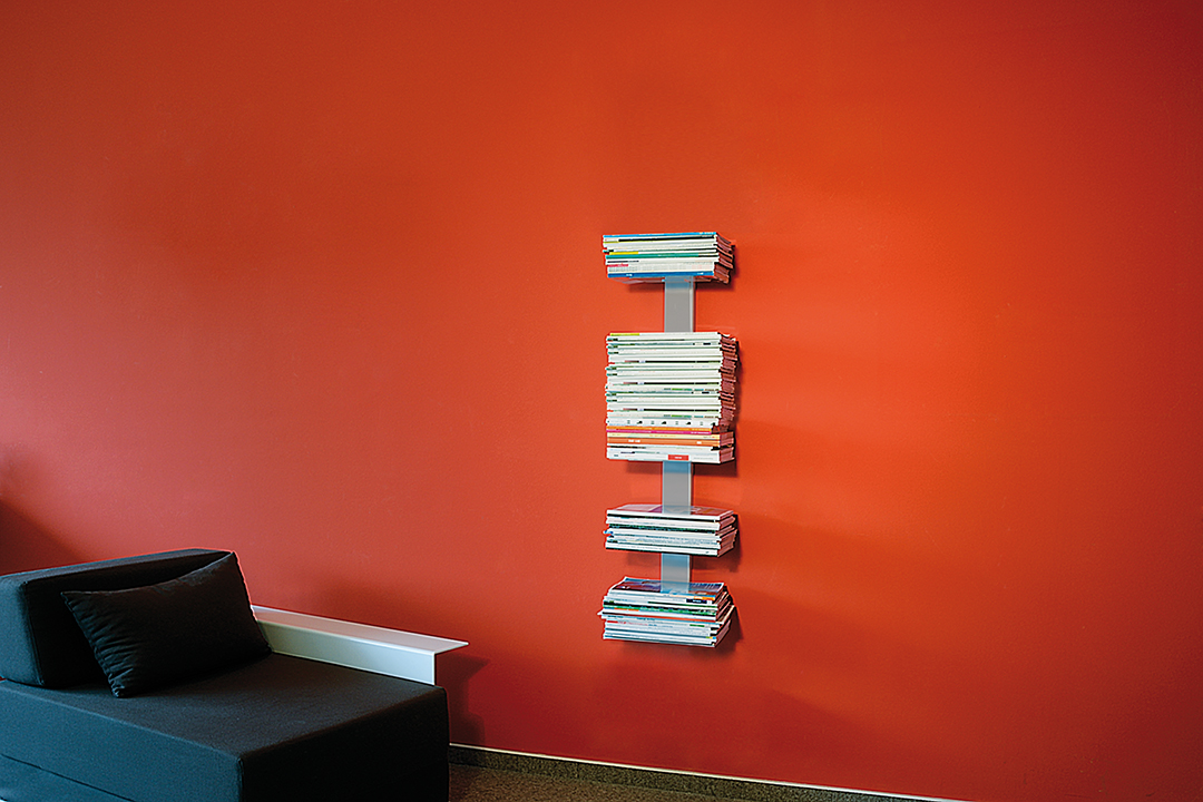 Booksbaum Wandregal mit Magazinen an roter Wand, daneben Sofa. Bücherregal für Wohnzimmer.