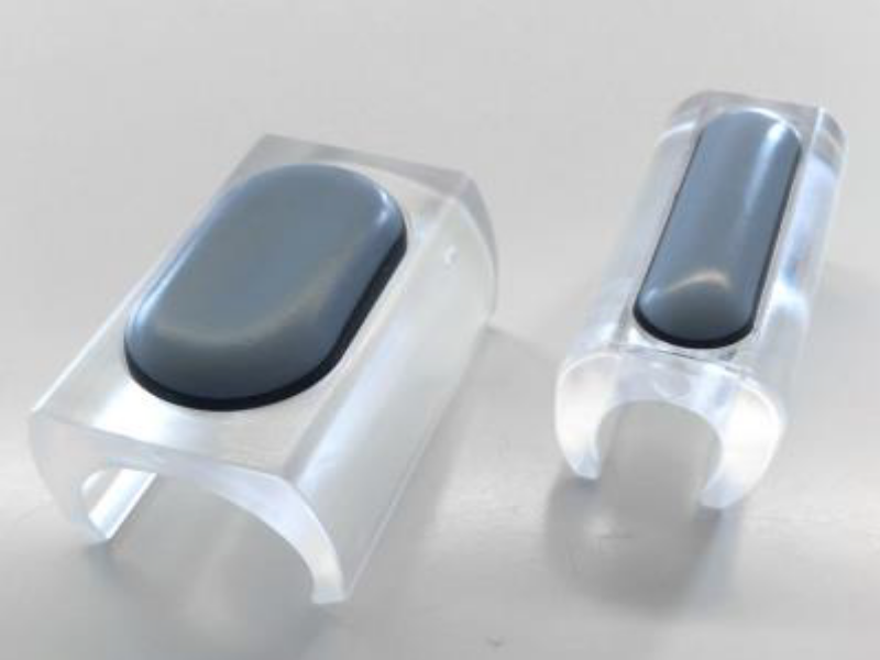 Zwei transparente Acryl-Kabelclips mit grauen Silikonpads zur Kabelführung und Organisation auf einem weißen Tisch.
