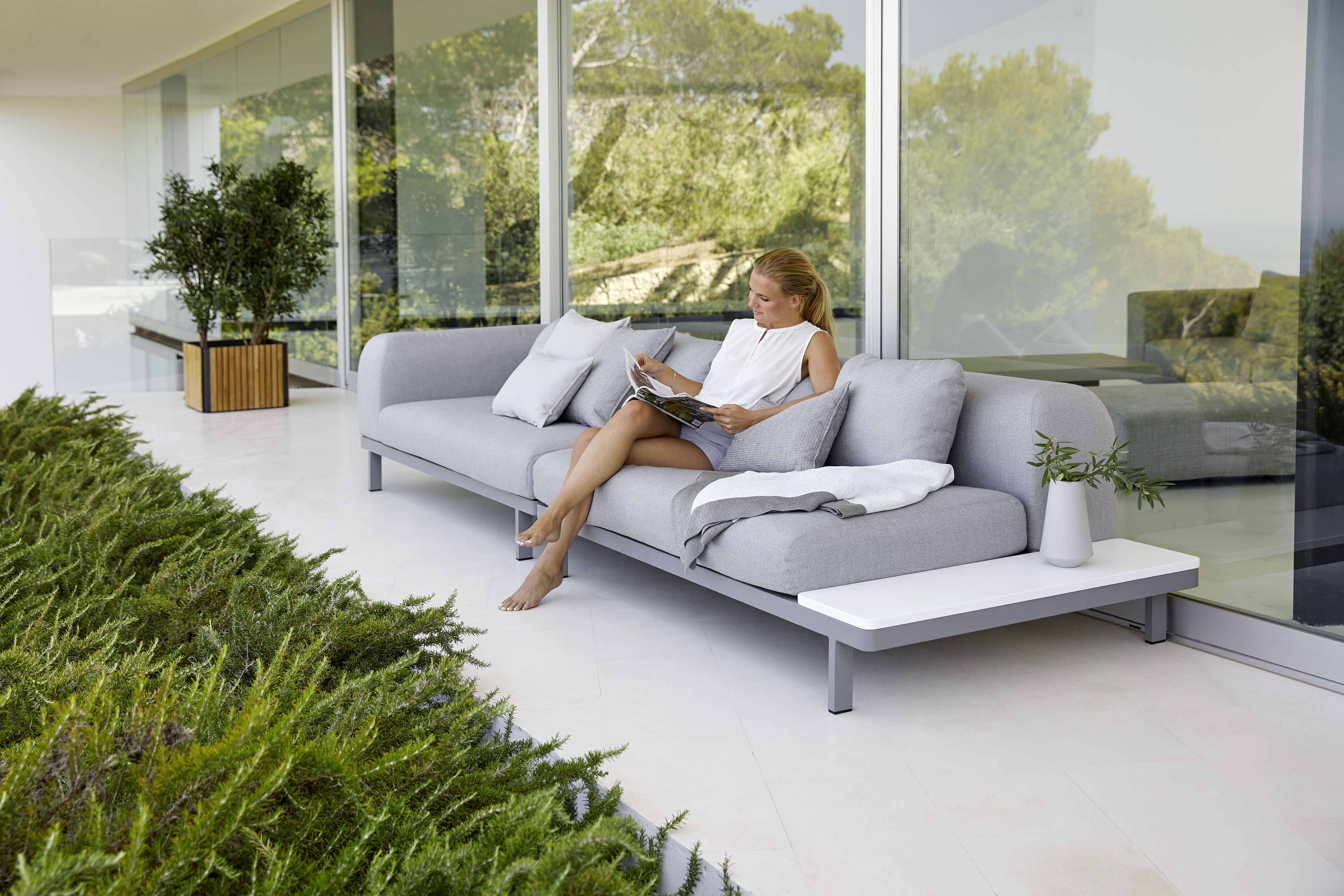 Frau liest auf einem grauen Cane-Line Sofa neben einem quadratischen Pflanzengefäß aus Holz im Garten.