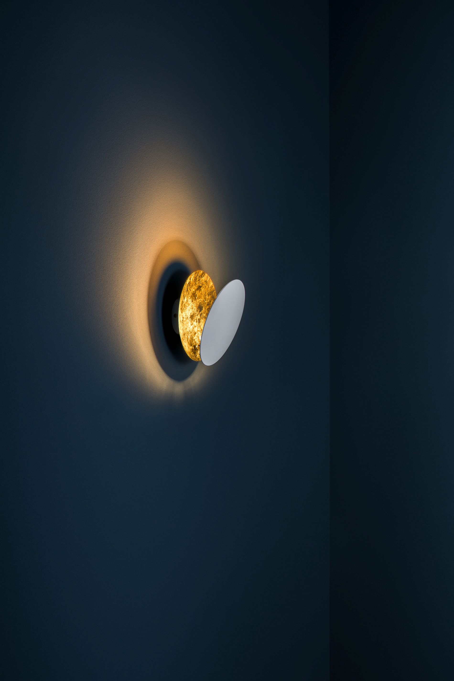Lederam Wandleuchte von Catellani & Smith: Goldene Scheibe mit weißem Reflektor an dunkler Wand.