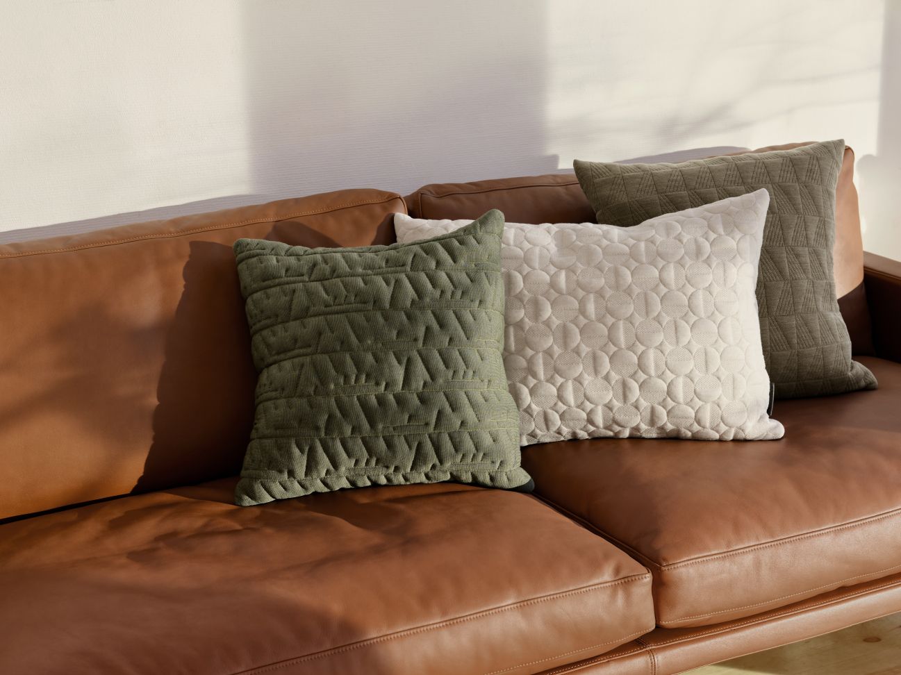Drei dekorative Kissen auf einem braunen Ledersofa, darunter ein Vertigo Arne Jacobsen Kissen in Sand.