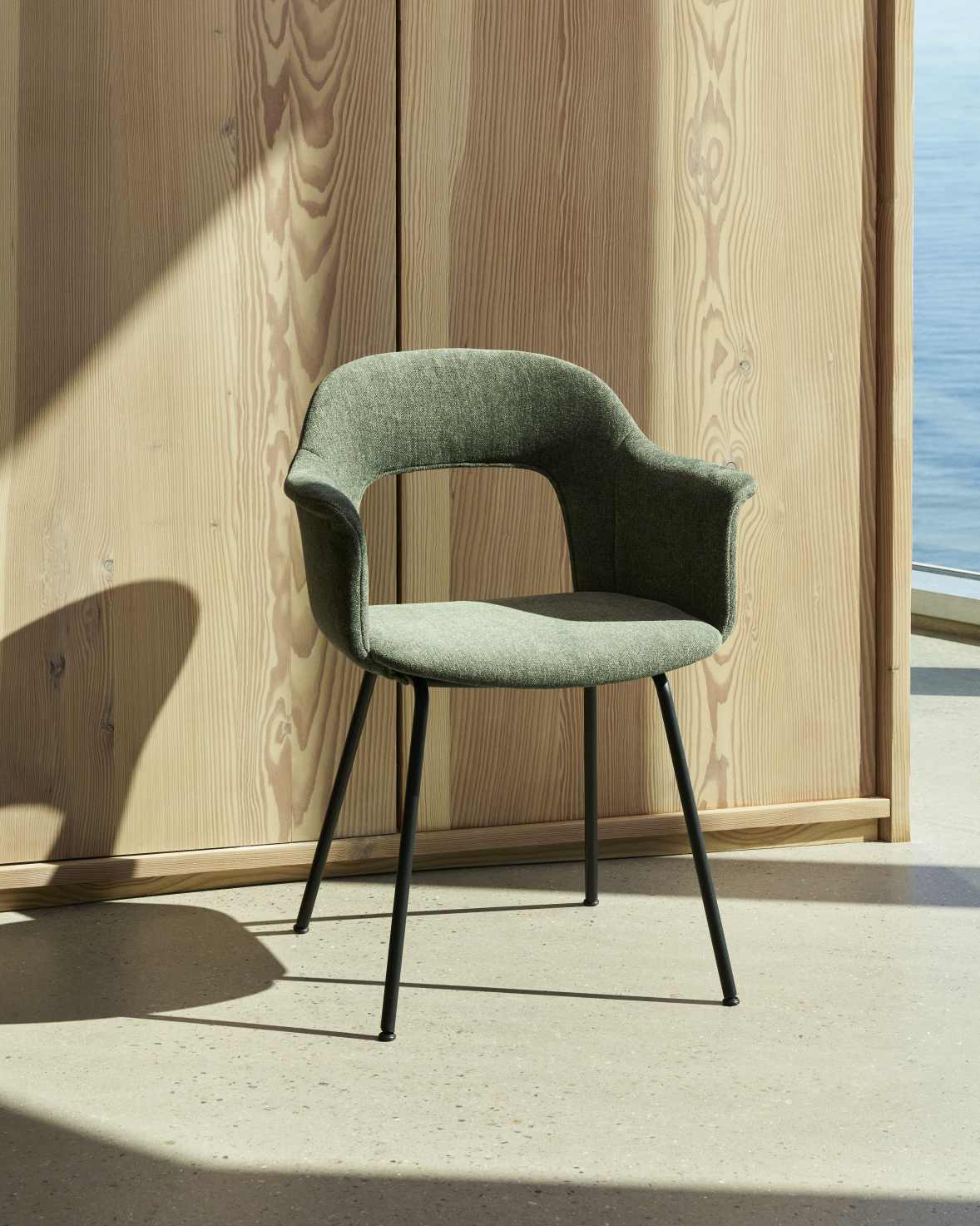 Layout Armchair 214 von Hay, grüner Armlehnstuhl mit schwarzem Metallgestell vor Holzwand und Meer.