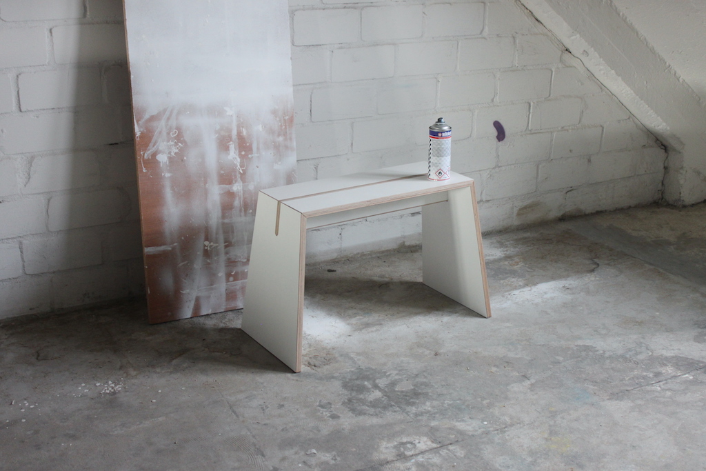 Tojo Stubenhocker Hocker aus Holz, weiß lackiert, mit Farbspraydose auf Betonboden vor Backsteinwand.