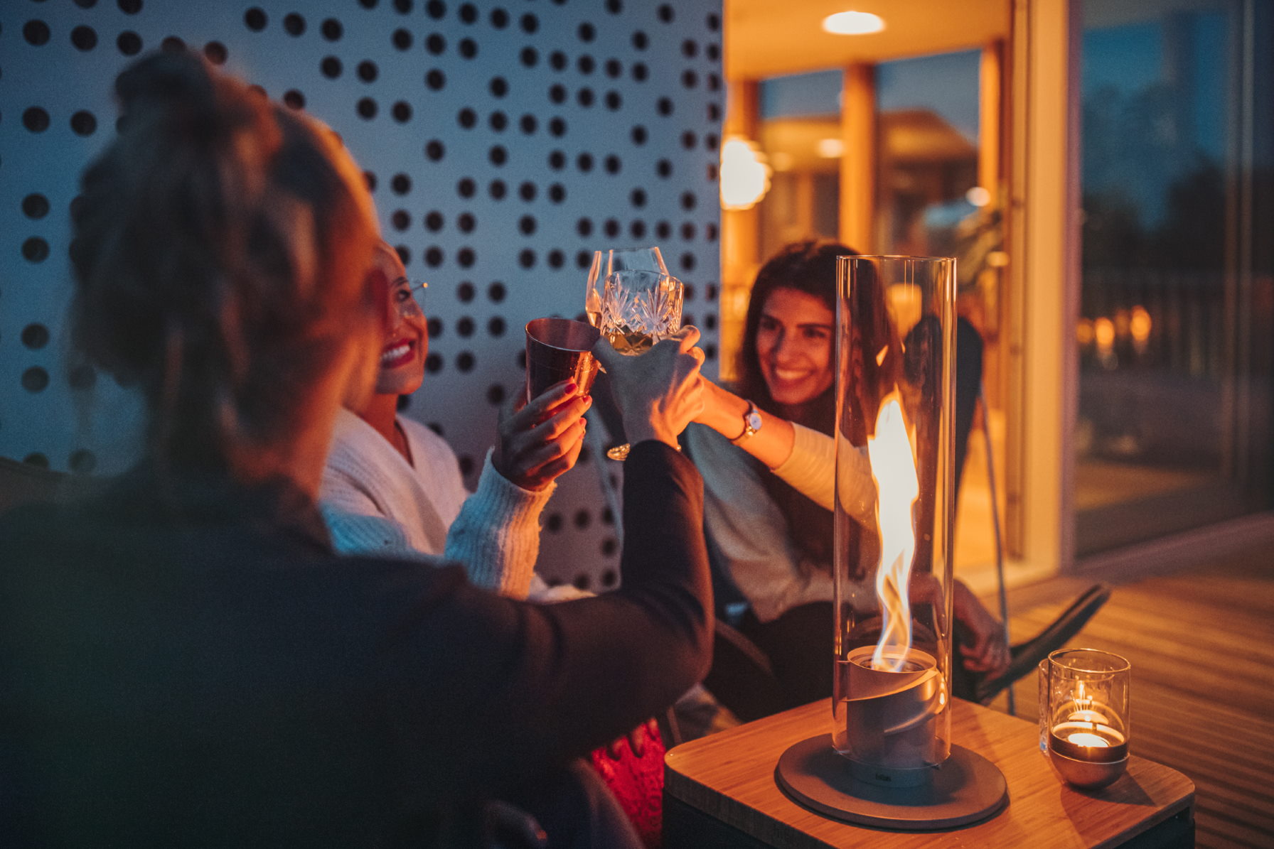Freunde stoßen mit Gläsern neben einem Tischkamin mit Glaszylinder im Freien an.