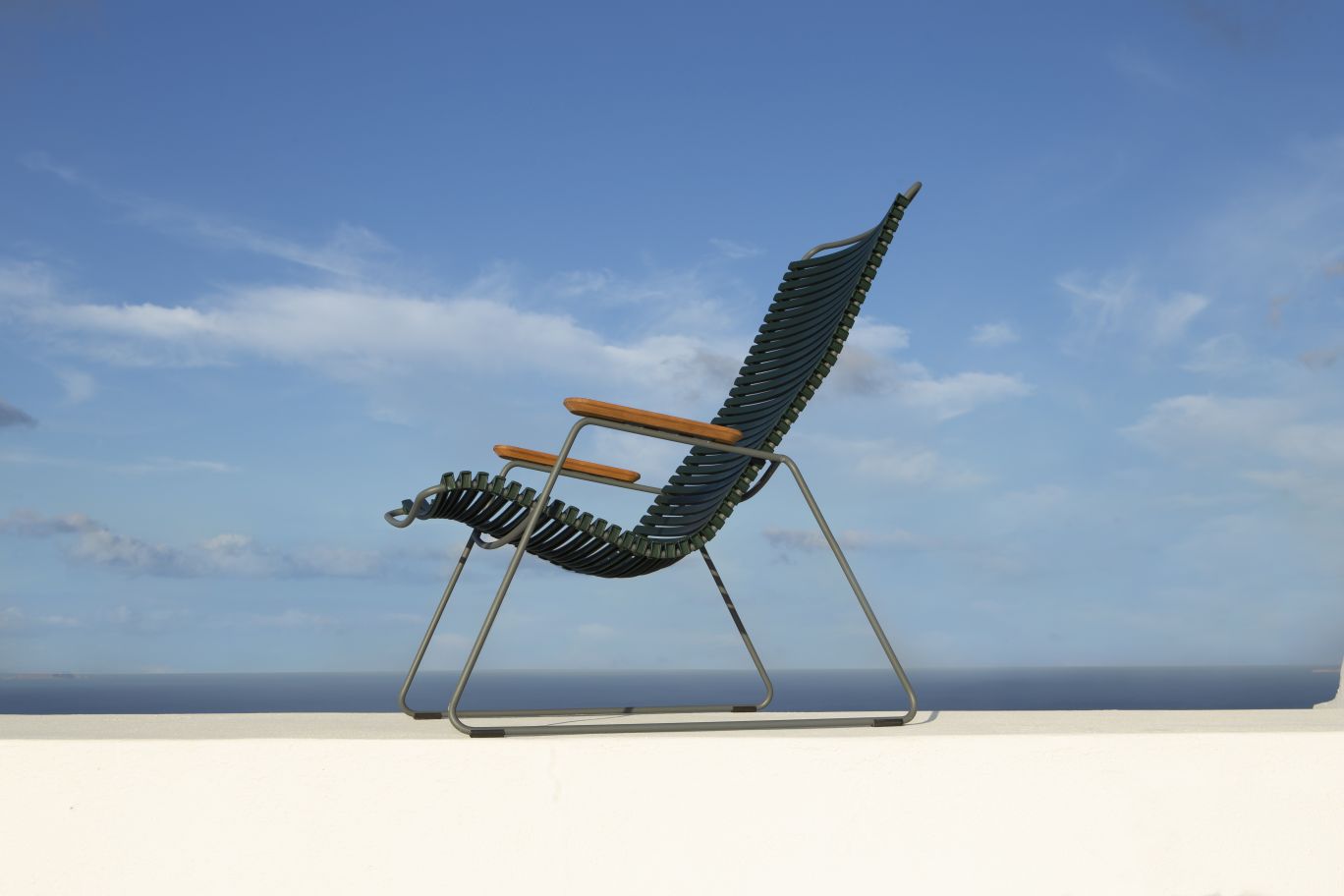 Houe Click Lounge Chair Outdoor Sessel mit Armlehnen vor blauem Himmel und Meer.