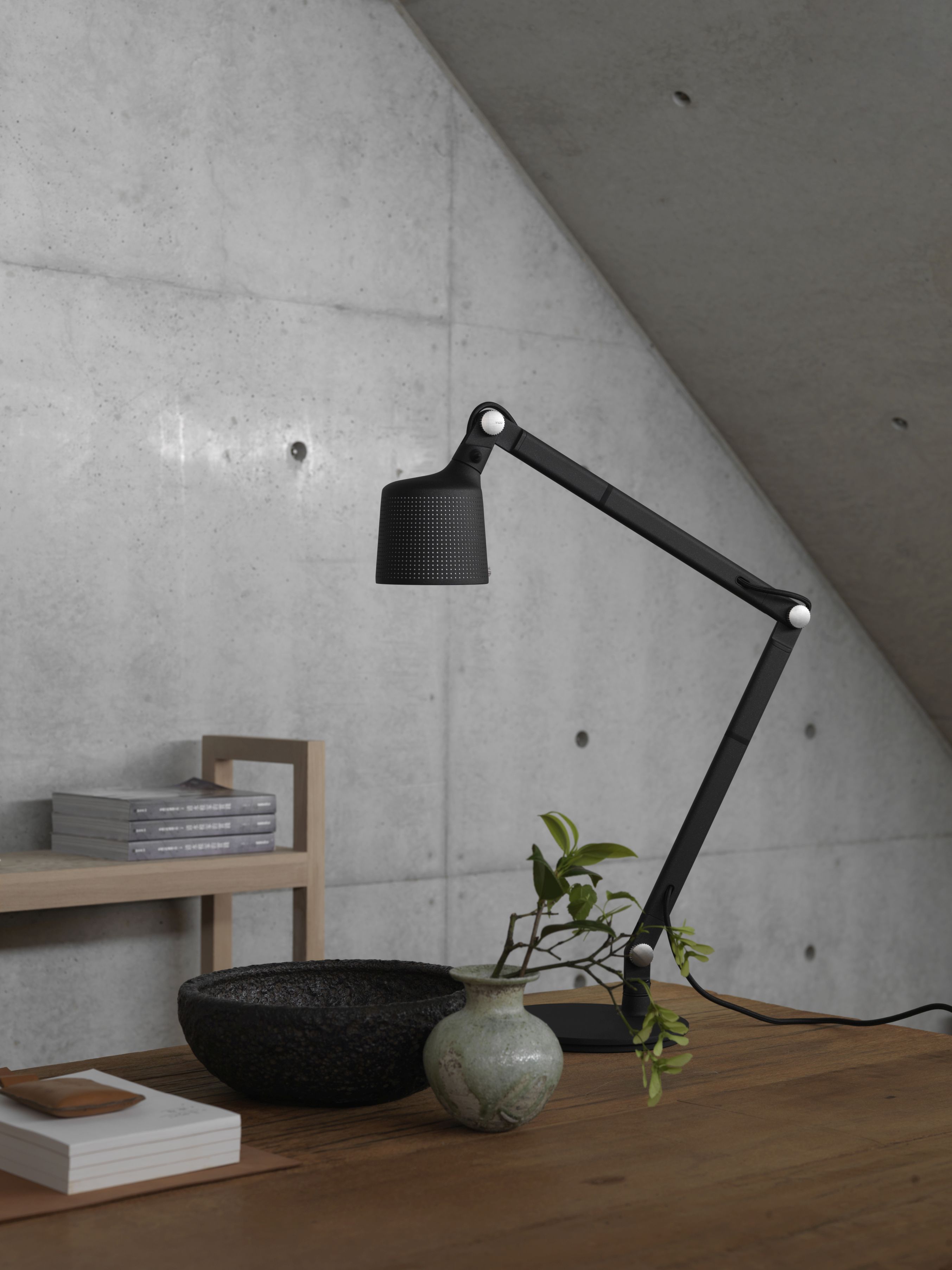 Schwarze Vipp Tischlampe auf einem Holztisch mit Deko-Elementen vor einer Betonwand.