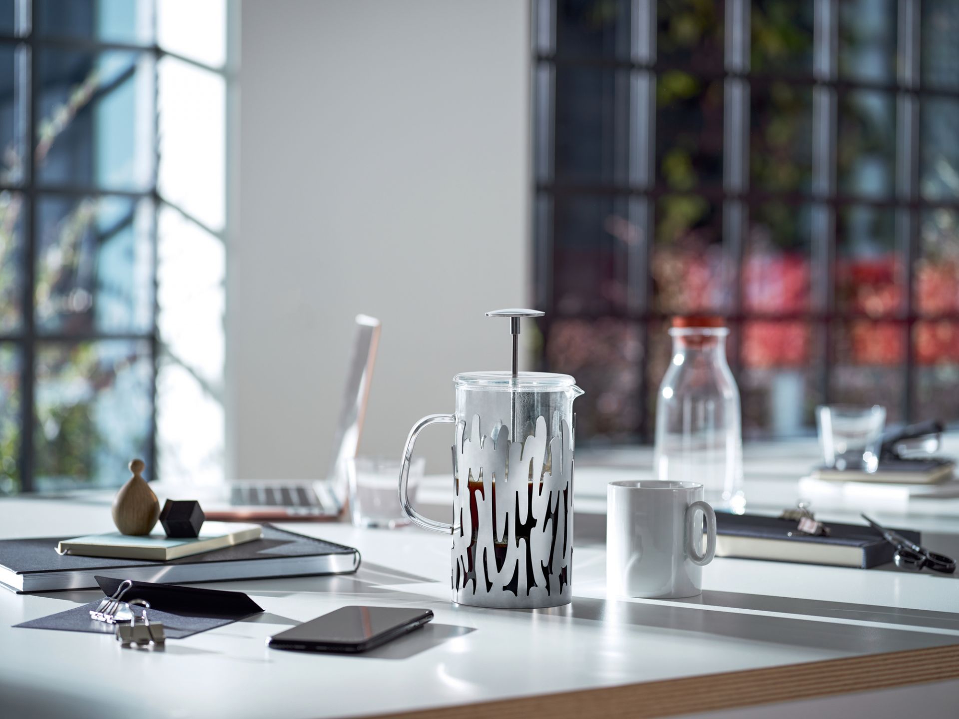 Moderne Glas-Kaffeepresse mit silberfarbenem Design und weißer Tasse auf einem Schreibtisch mit Büromaterial.