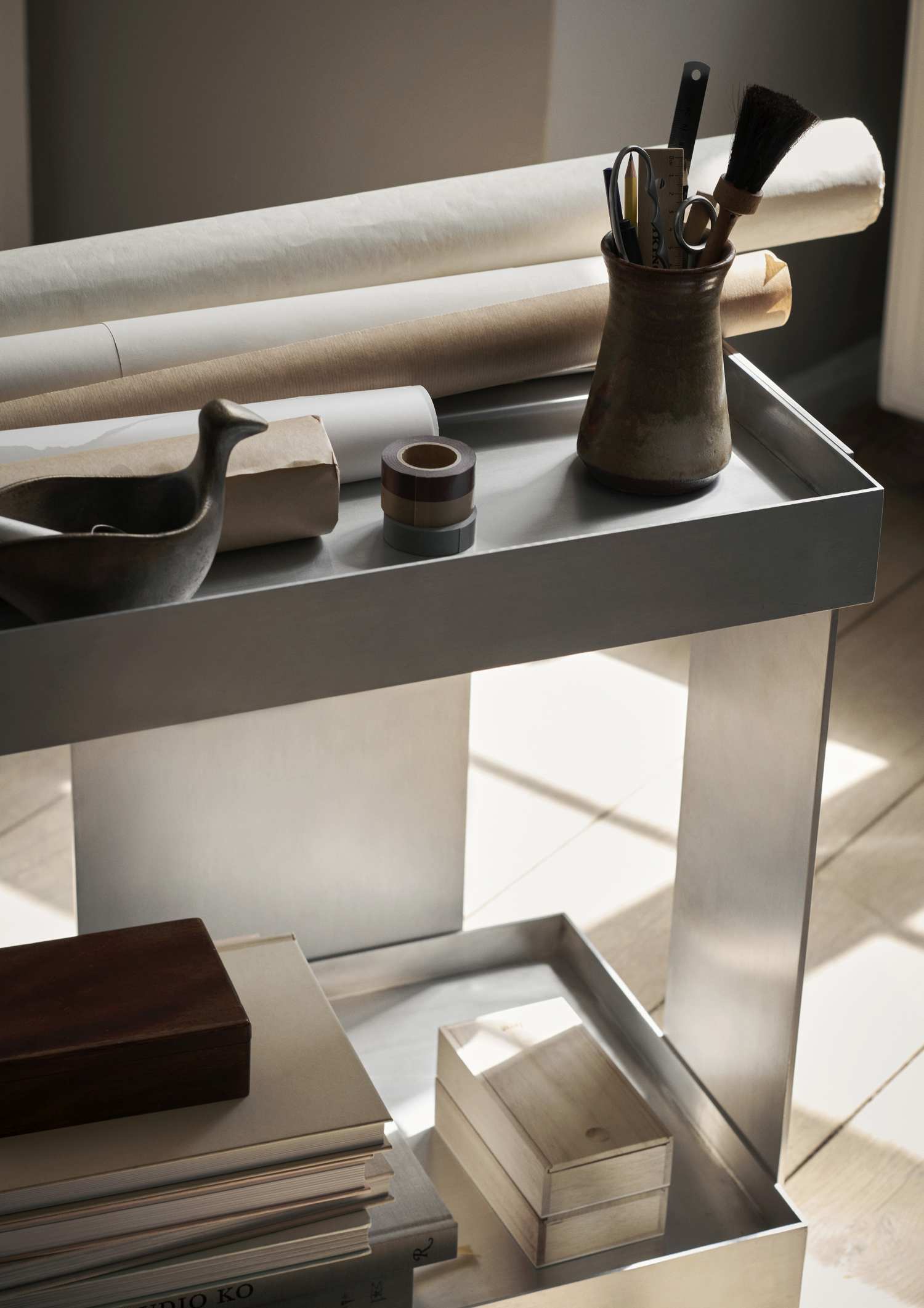 Ferm Living Lager Trolley: Moderner Aluminium Servierwagen mit zwei Ebenen, dekoriert mit Büroartikeln und Papieren.