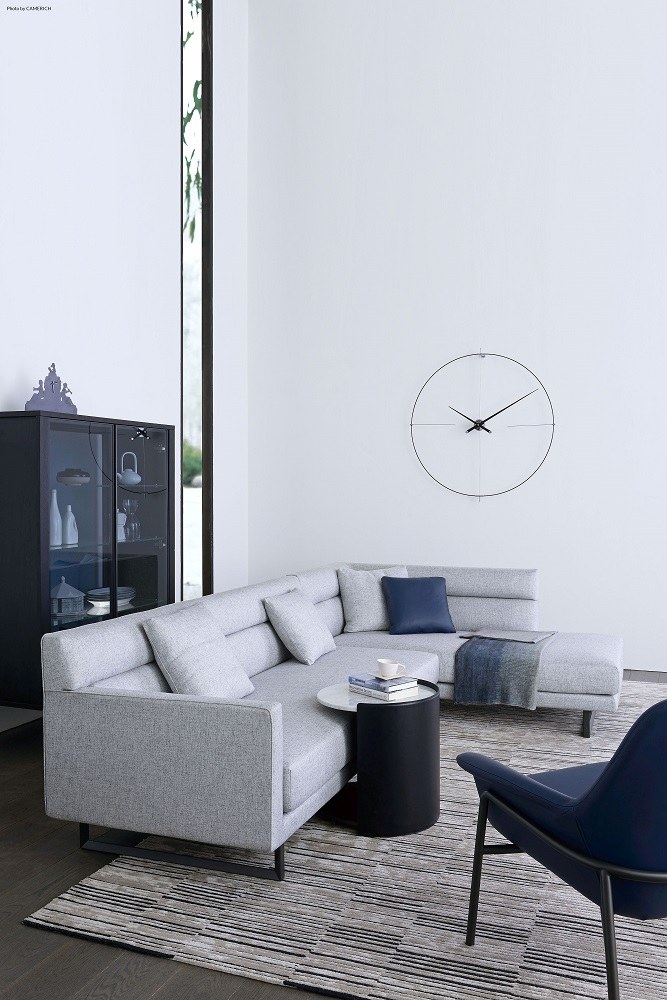 Wohnzimmer mit grauer Eckcouch, schwarzem Beistelltisch und Bilbao Nomon Wanduhr in Schwarz.