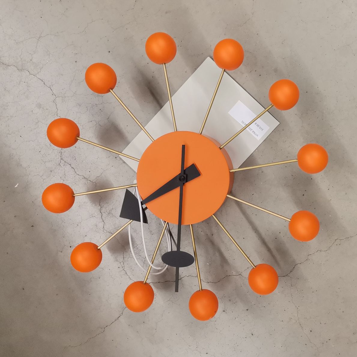 Orangefarbene Ball Clock Wanduhr von Vitra mit goldenen Streben und schwarzen Zeigern auf grauem Hintergrund.
