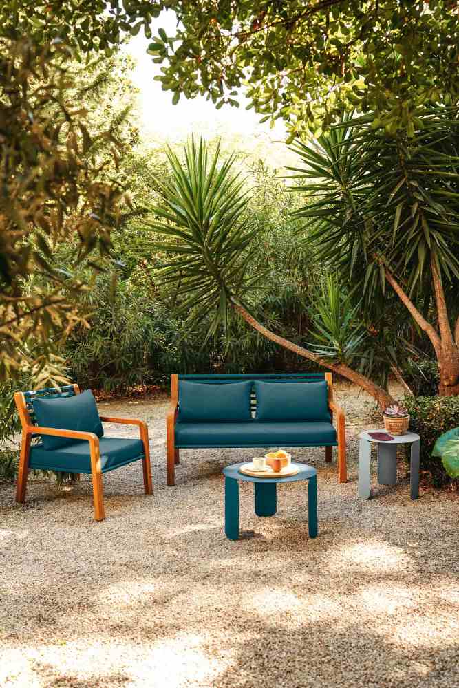 Somerset 2-Sitzer Sofa von Fermob mit Sessel und Tisch in einem grünen Garten.