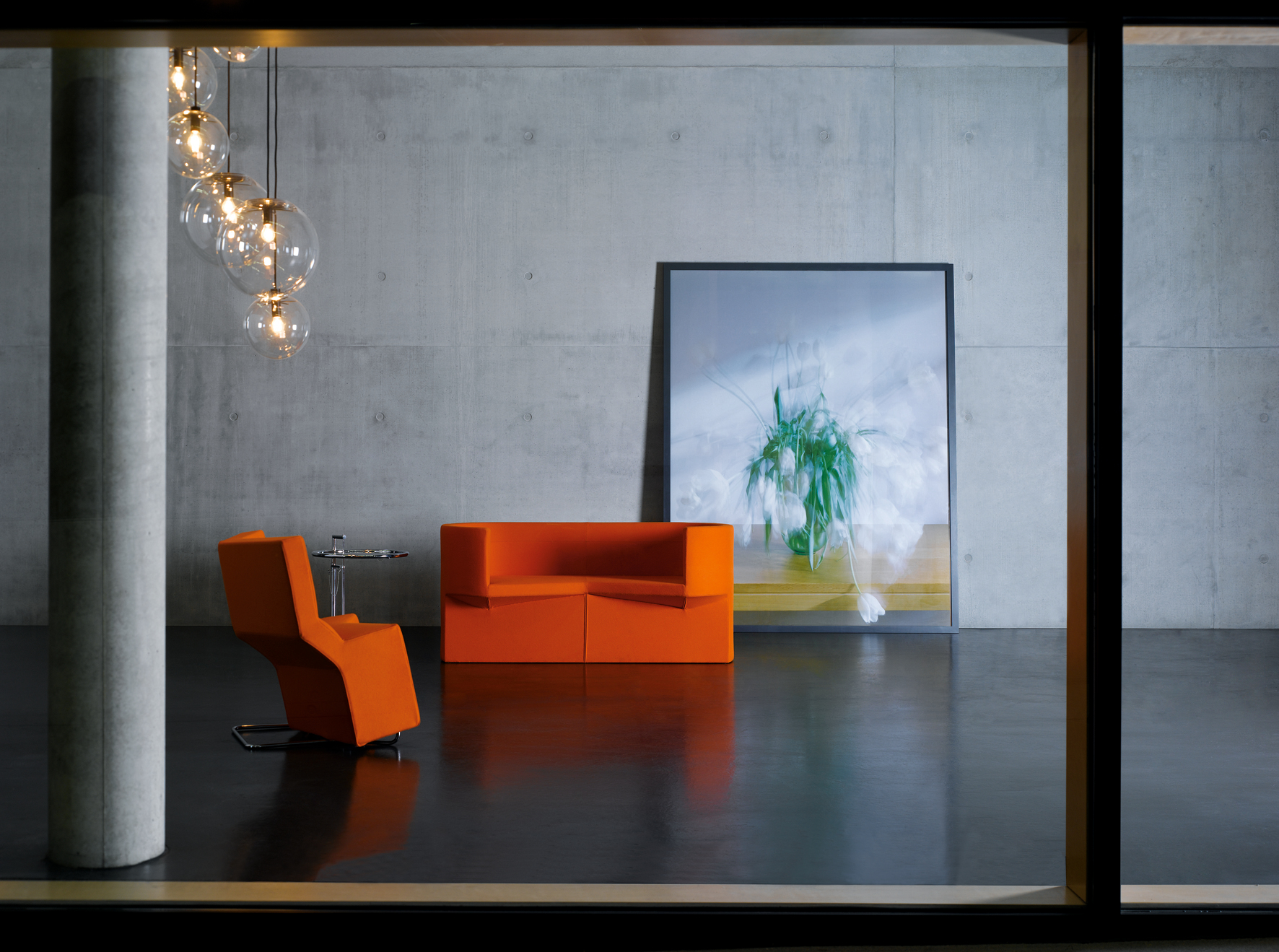 Moderner Wohnraum mit orangefarbenem Sessel, Sofa und ClassiCon E1027 Beistelltisch mit Glasplatte vor Betonwand.