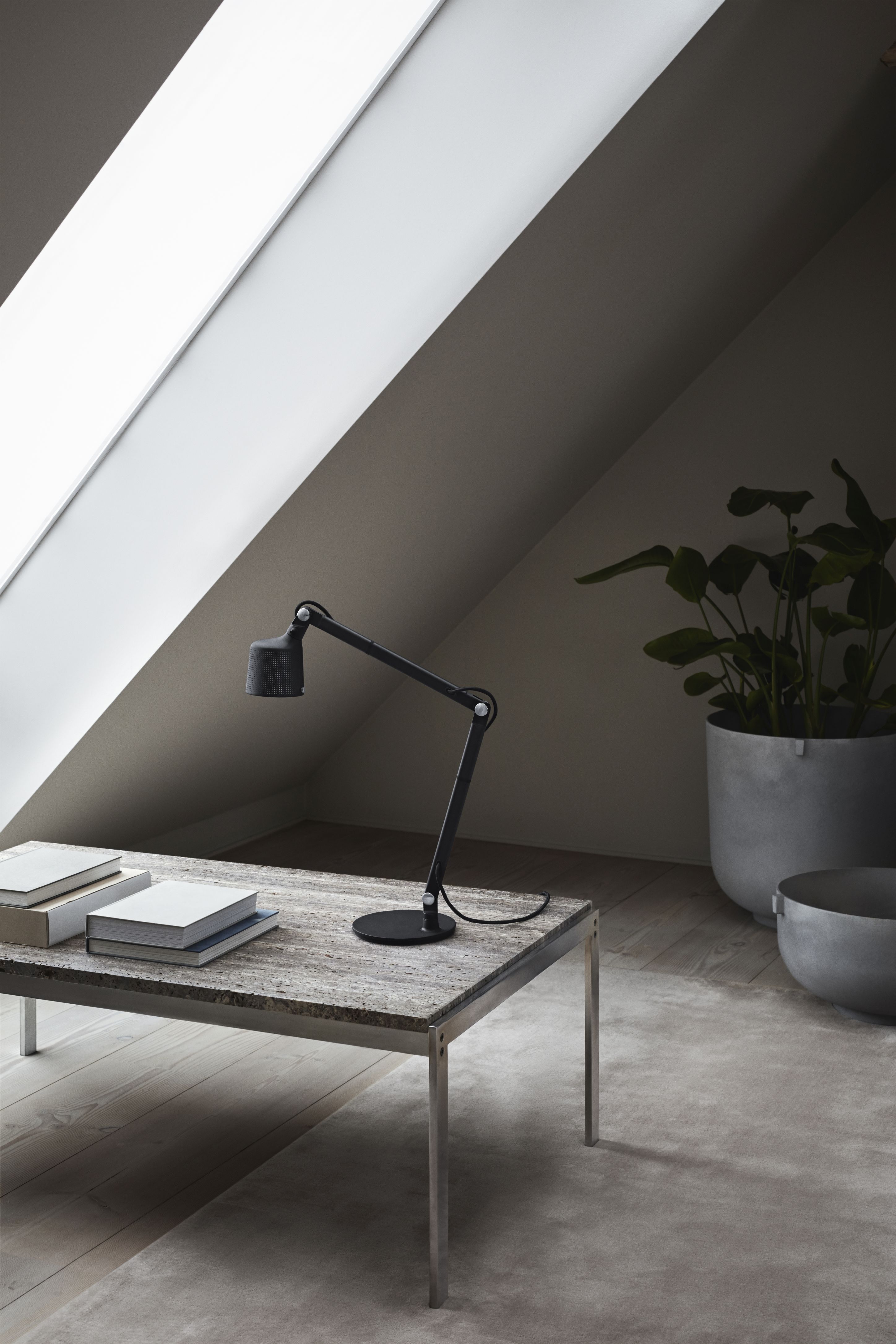 Schwarze Vipp Tischlampe auf Holztisch mit Büchern, Zimmerpflanze und Dachfenster im Hintergrund.