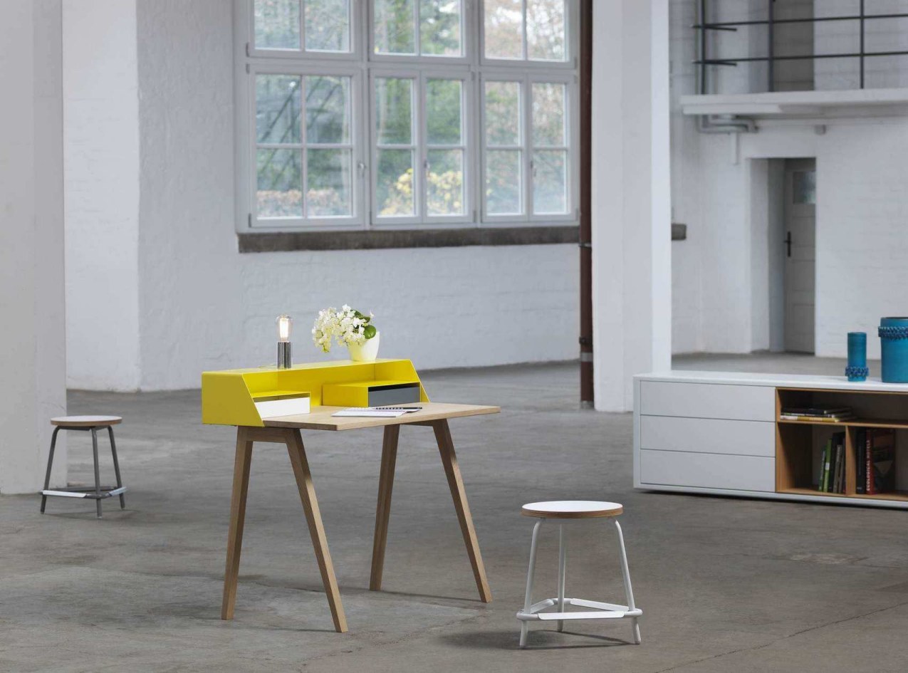 Gelber Schreibtisch mit Holzbeinen, Lampe und Blumen in hellem Raum. Moderner Bürotisch für Homeoffice.