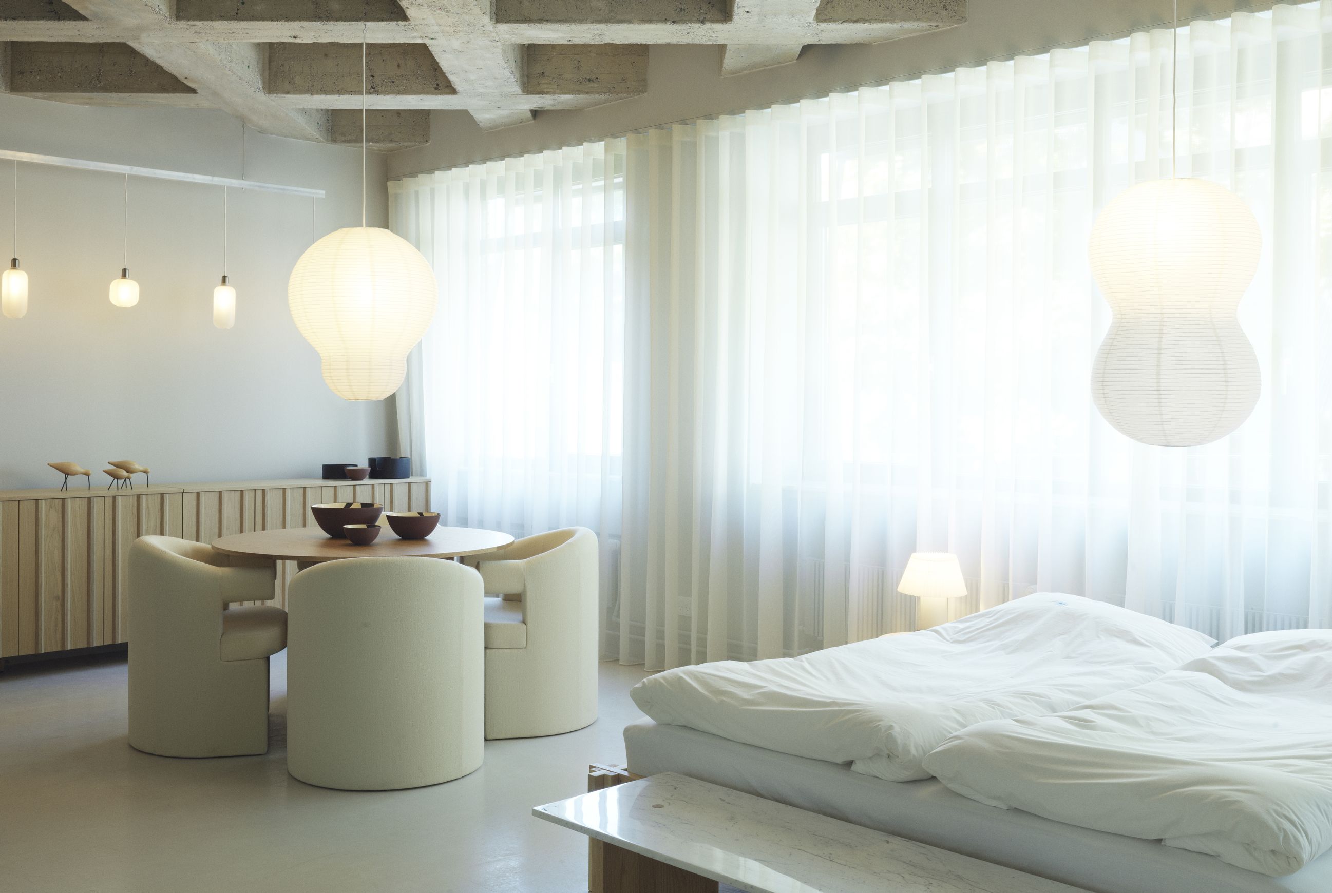 Helles Interieur mit Normann Copenhagen Burra Stuhl, Holztisch, Lampen und Bett mit weißer Bettwäsche.