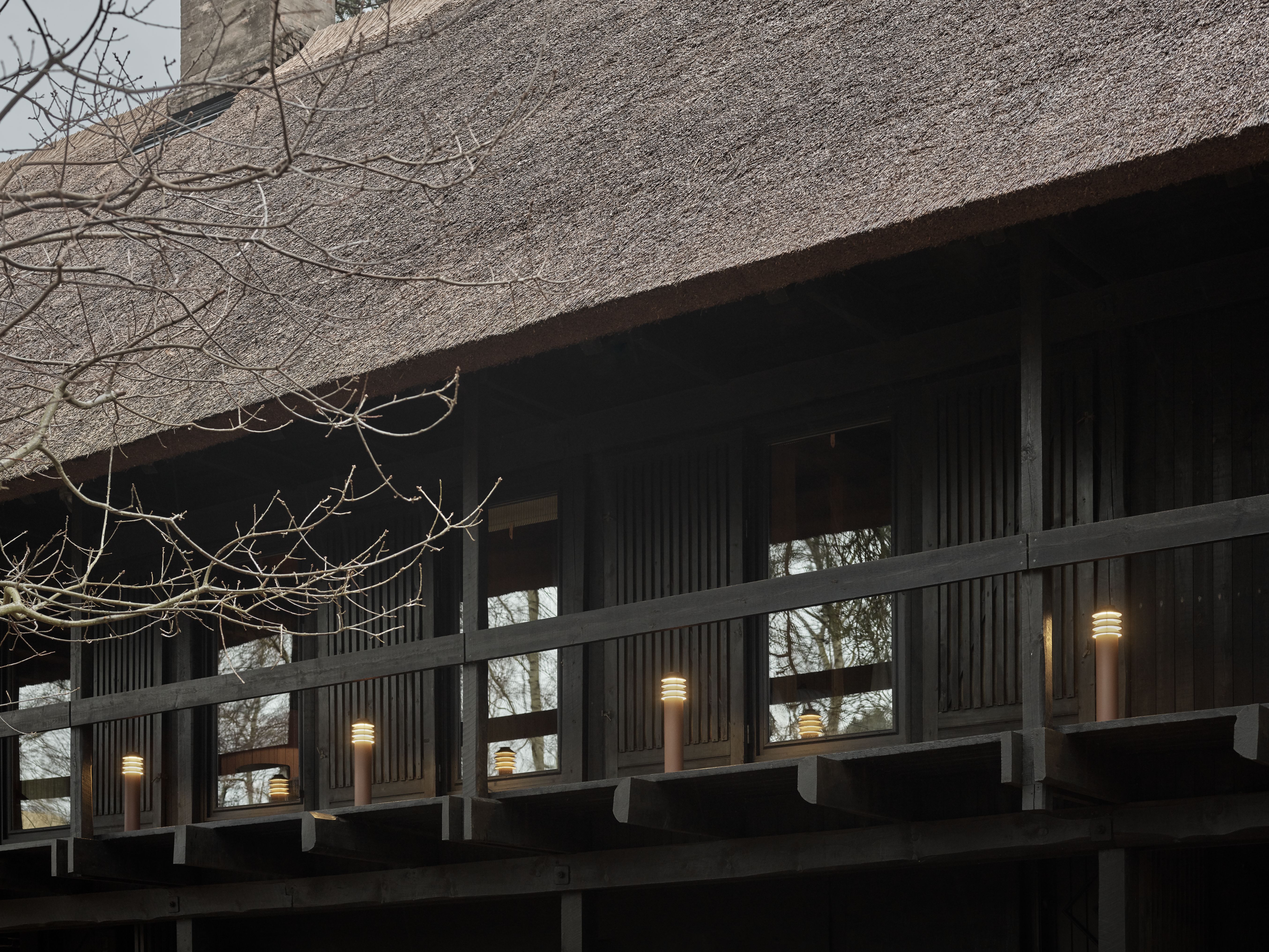 Nahaufnahme der Louis Poulsen Bysted Garden Pollerleuchten vor einem dunklen Holzhaus mit Reetdach.