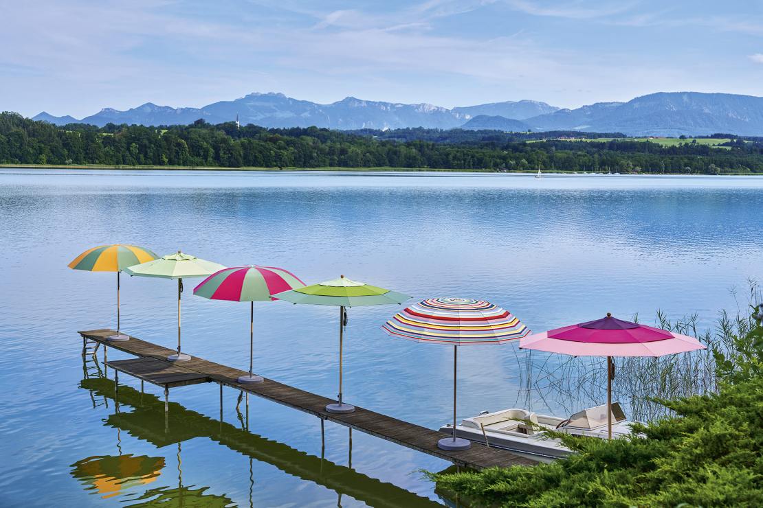Mehrere farbenfrohe Weishäupl Sonnenschirme/Pagodenschirme stehen auf einem Holzsteg an einem See mit Bergen im Hintergrund.