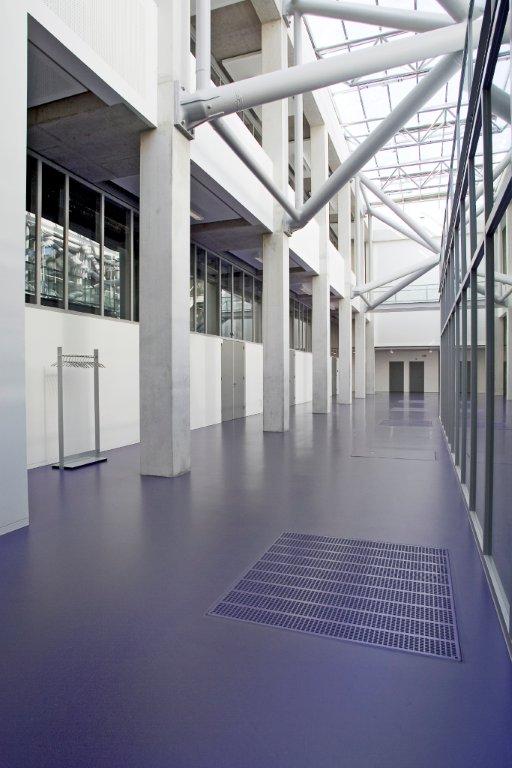 Helle Industriehalle mit lila Epoxidharzboden, Metallgitter, Fensterfronten und Stahlträger-Deckenkonstruktion für moderne Architektur und Design.