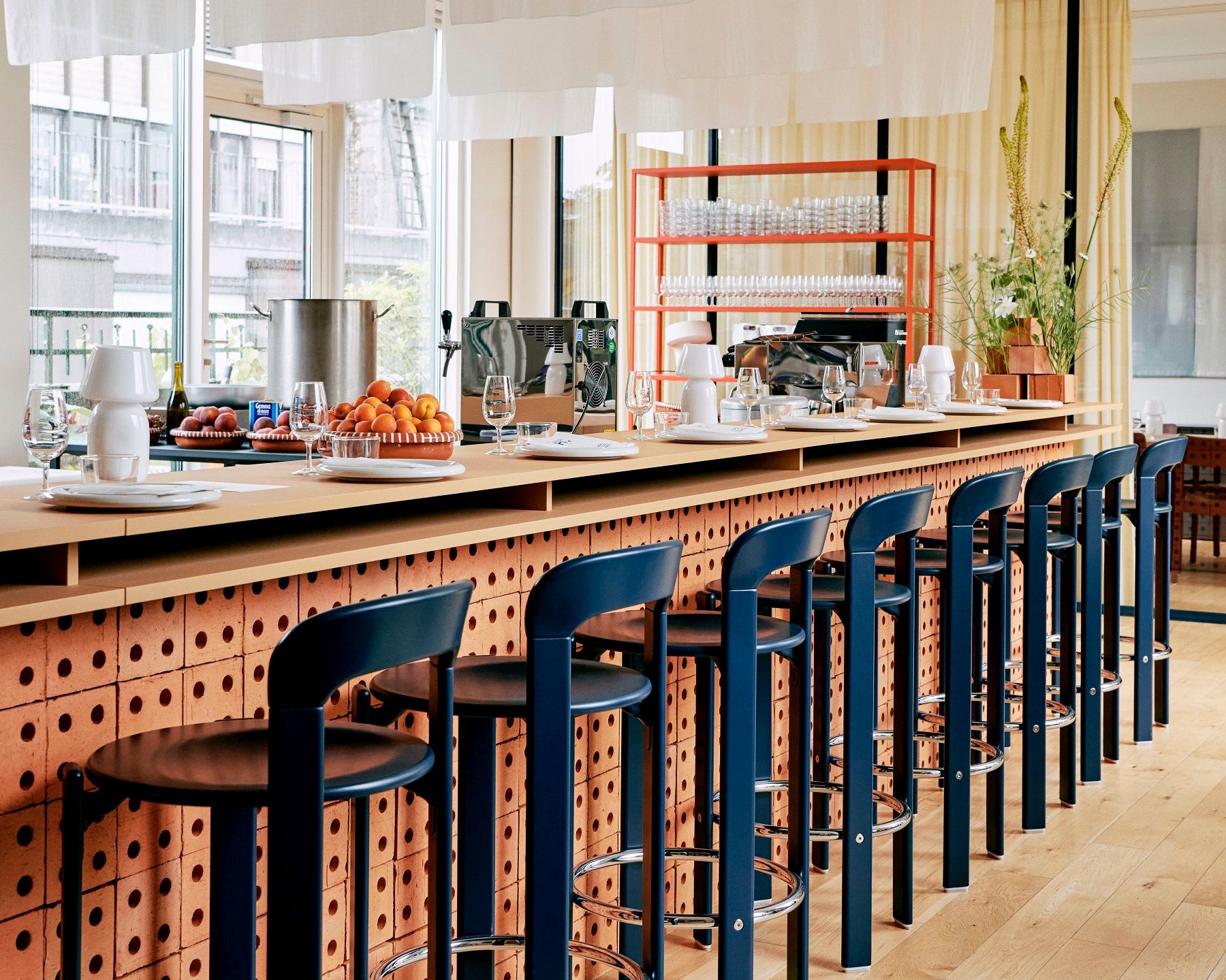 Reihe von blauen Rey Barhocker H97cm von Hay an einer Bar mit Backsteinmuster.