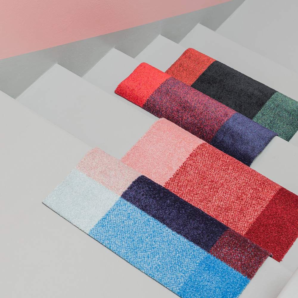 Mehrere farbenfrohe Heymat Fußmatten für Innen und Außen auf einer grauen Treppe, verschiedene Designs.