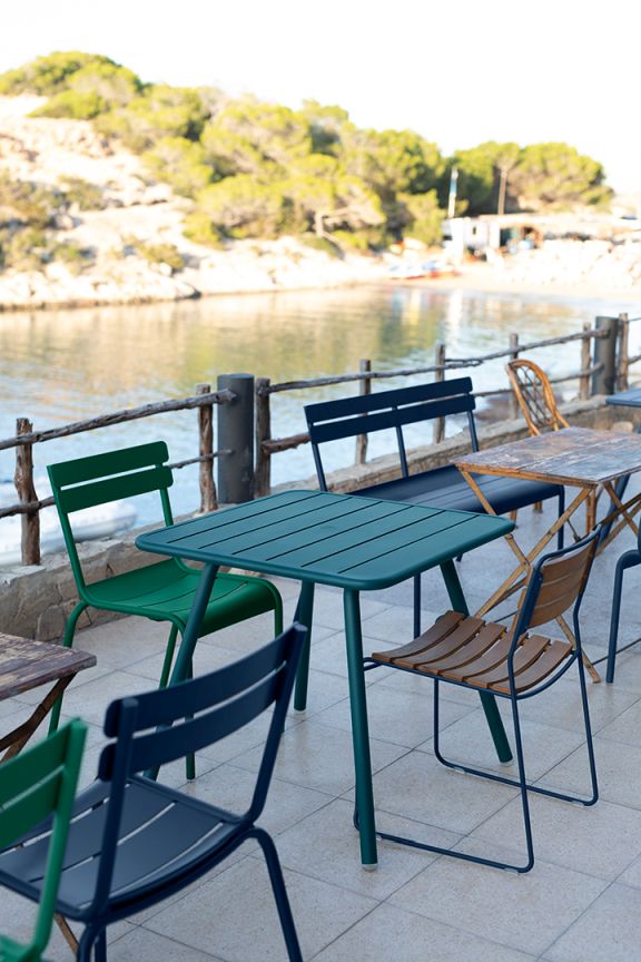 Nahaufnahme von Fermob Luxembourg Outdoor Stahlstühlen und Tisch in Grün und Blau vor Küstenkulisse.