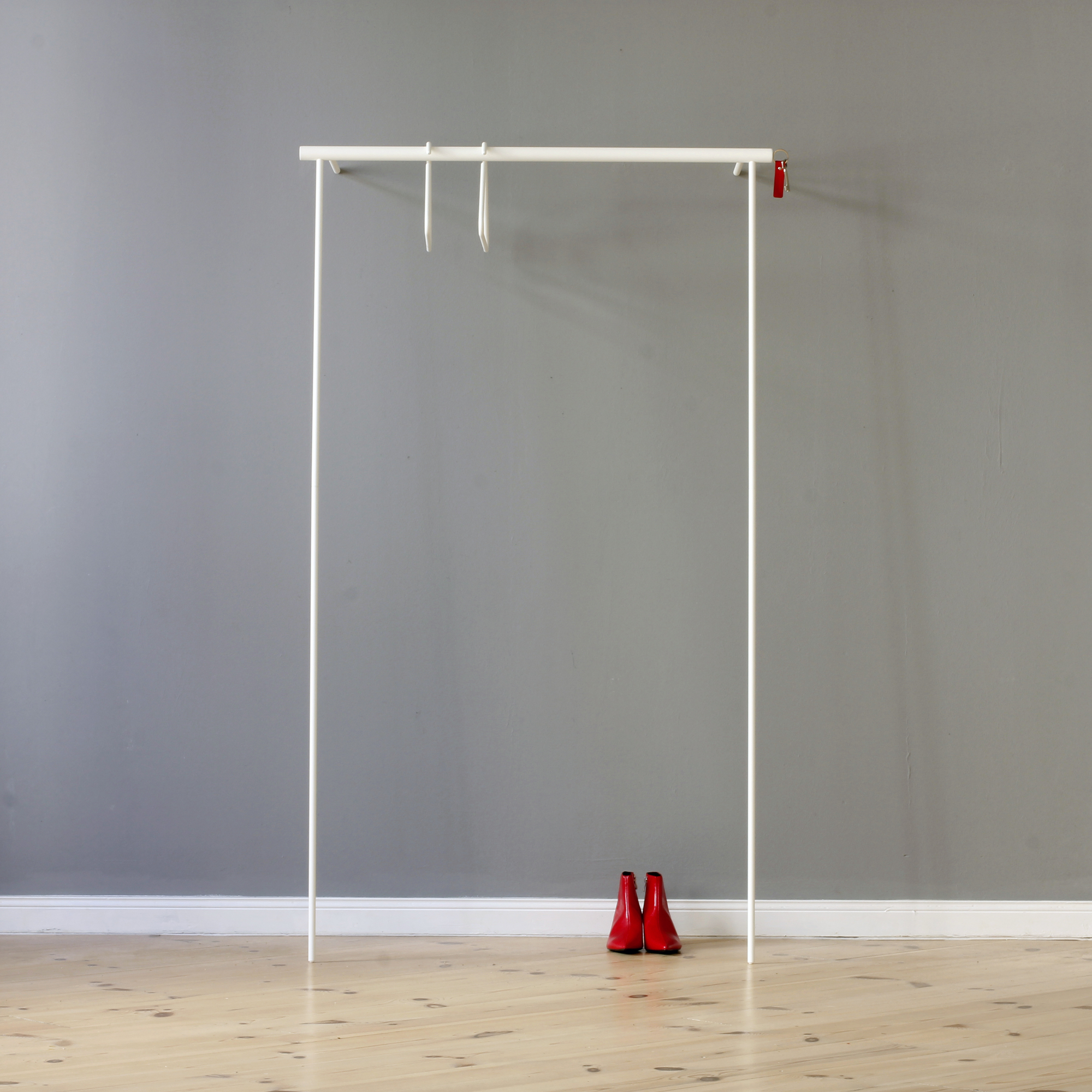 Leanon II Anlehngarderobe von Roomsafari, weiße minimalistische Kleiderstange mit roten Schuhen davor, vor grauer Wand.