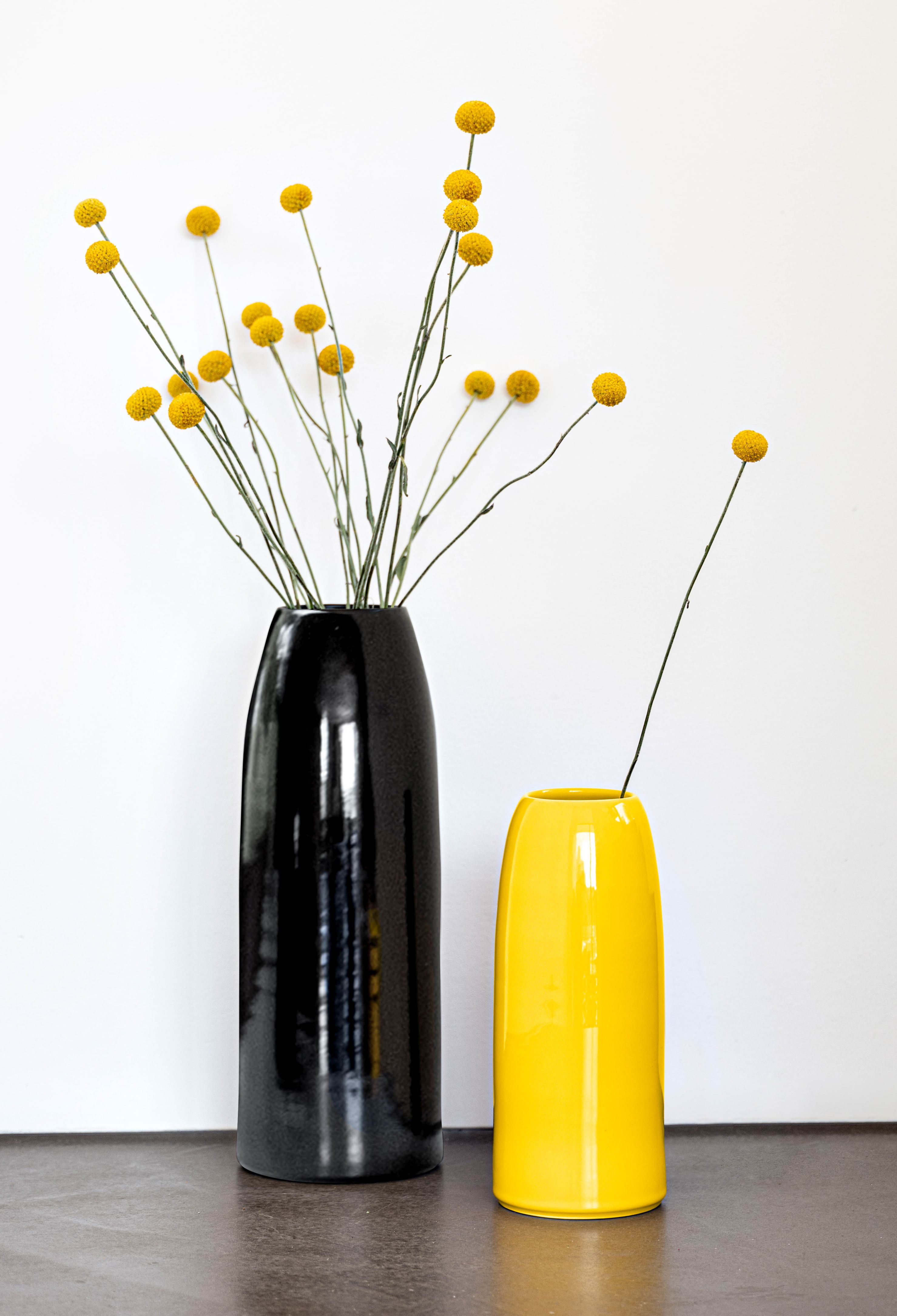 Schönbuch Carla Vase L: Schwarze Design-Vase und gelbe Vase mit gelben Zierblumen auf einem Tisch.