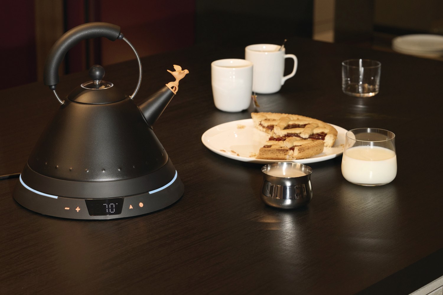 Schwarzer Alessi MG32 Wasserkocher mit Temperaturregelung, Vogel-Pfeife, Tasse Kaffee, Milch und Kuchen auf dunklem Tisch.