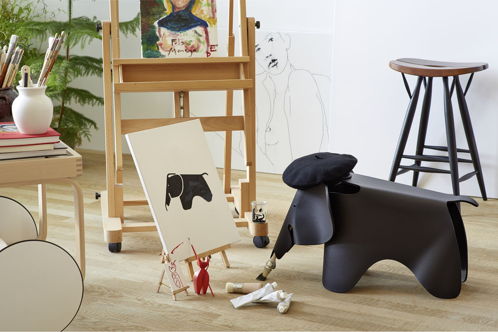 Schwarzer Eames Elefant Hocker mit Baskenmütze in einem Künstleratelier mit Staffelei und Malutensilien.