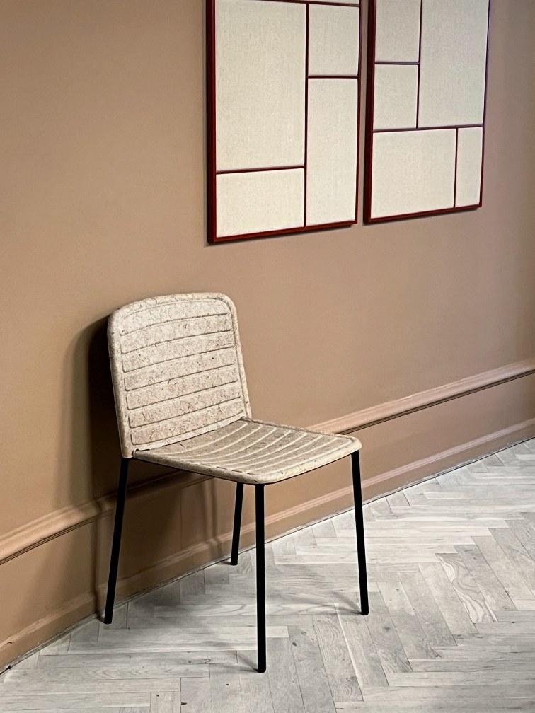 Flax Stacker Chair: Moderner Stuhl mit Leinenstruktur und schwarzen Beinen vor brauner Wand.