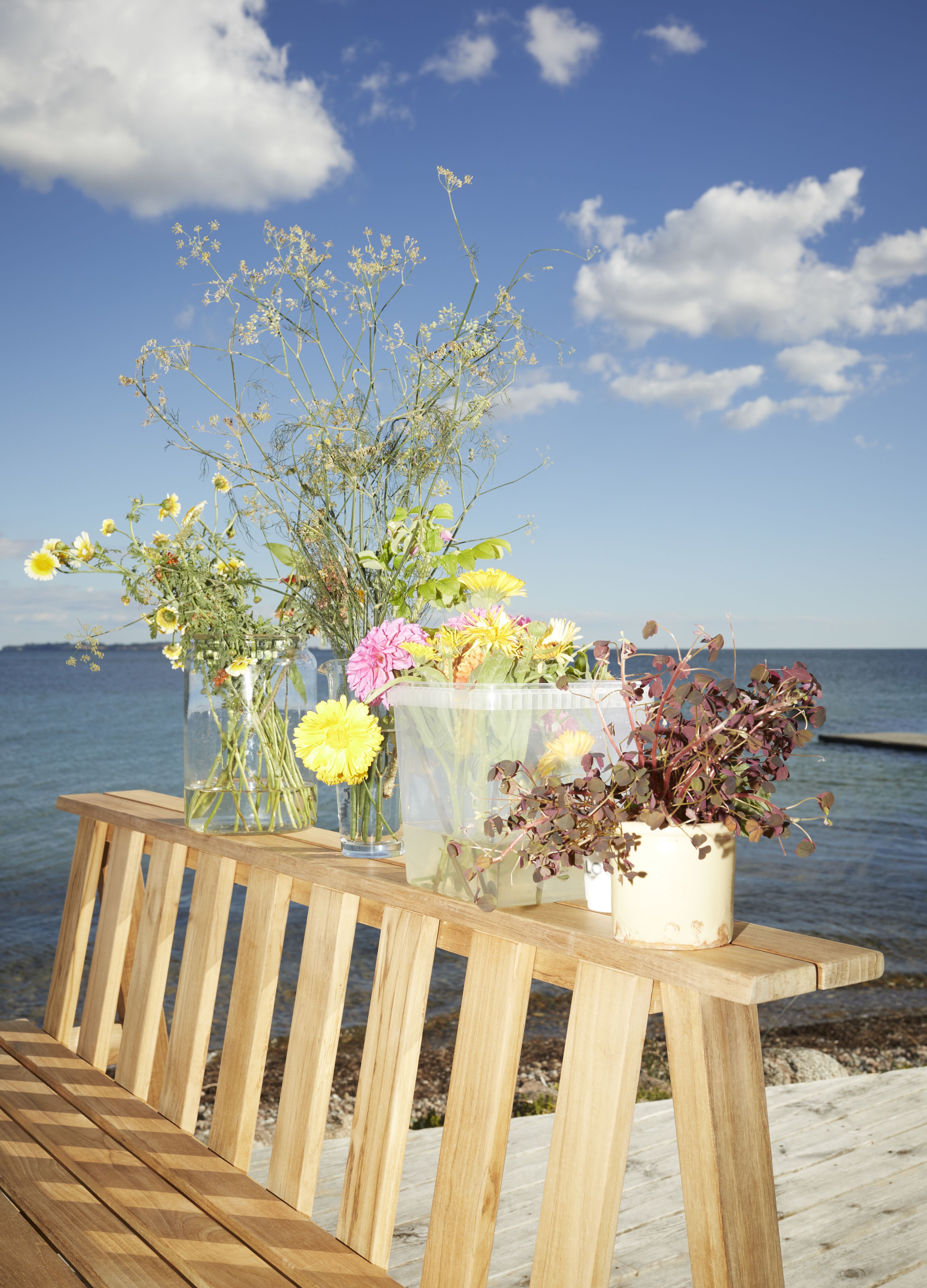 Cadence Bank Teak Outdoor Bank mit Blumenvasen und Topfpflanze vor blauem Himmel und Meer.