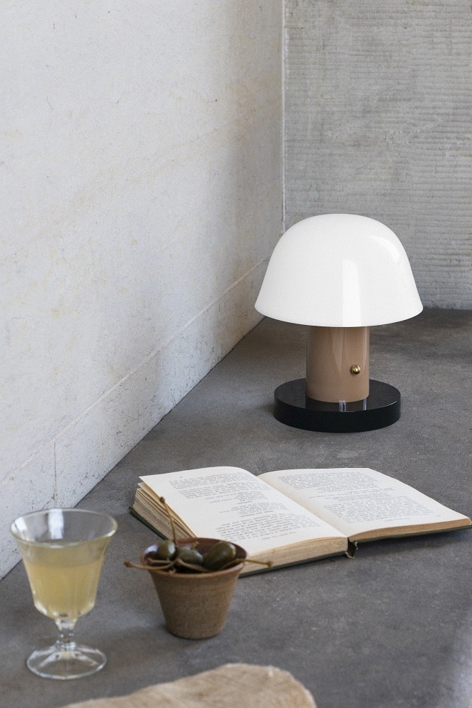 AndTradition Setago Tischleuchte JH27, weiße Pilzlampe neben Buch, Getränk und Kapern auf grauem Untergrund.