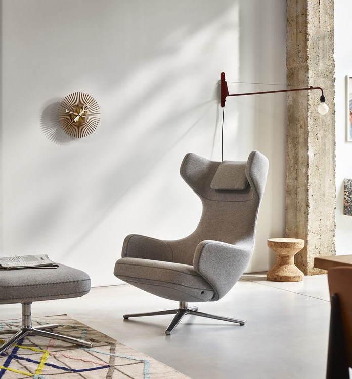 Grauer Ohrensessel mit Fußhocker und Metallfuß in modernem Wohnzimmer mit Lampe und Uhr.