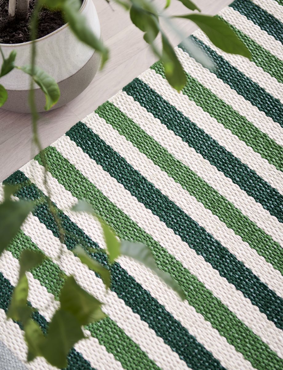 Detailaufnahme des Teo Teppichs von Pappelina (230x320cm) mit grün-weißem Streifenmuster und Pflanze im Hintergrund.