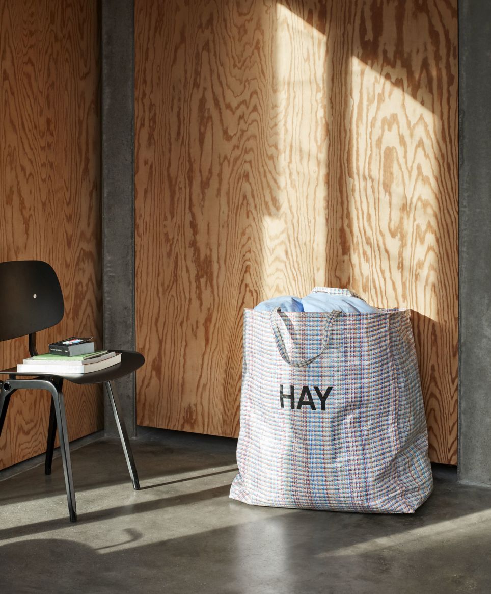 Schwarzer Hay Revolt Chair Stuhl mit Bücherstapel, daneben eine große Hay Einkaufstasche vor einer Holzwand.