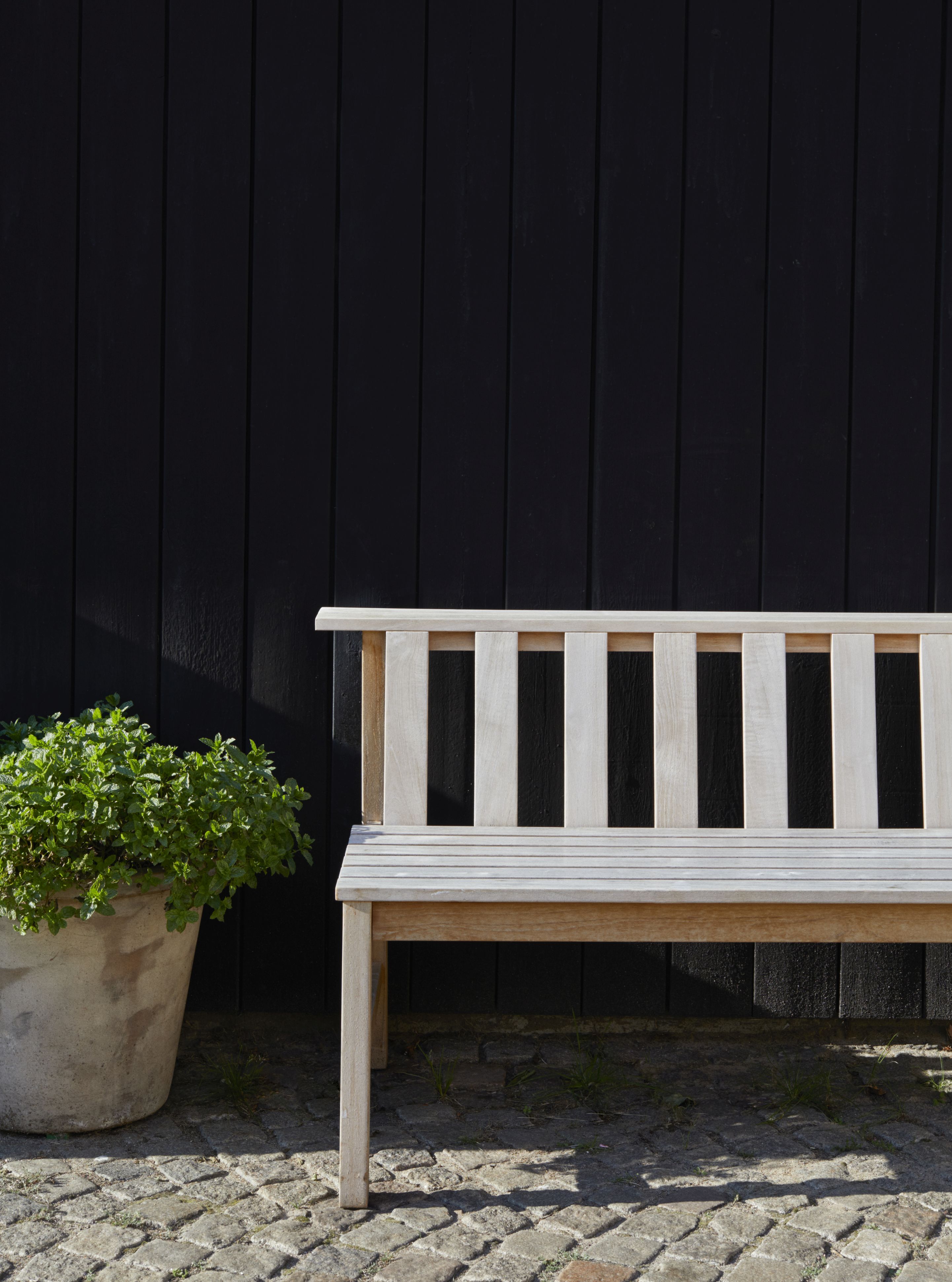 Cadence Bank Teakholz Gartenbank von Skagerak by Fritz Hansen vor schwarzer Holzwand und Pflanzkübel.