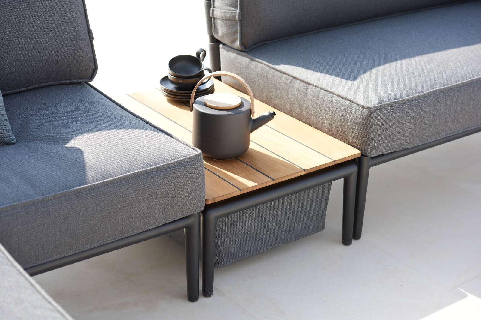 Cane-Line Conic Outdoor Box Tisch aus Teakholz mit Teekanne und Tassen zwischen grauen Loungemöbeln.