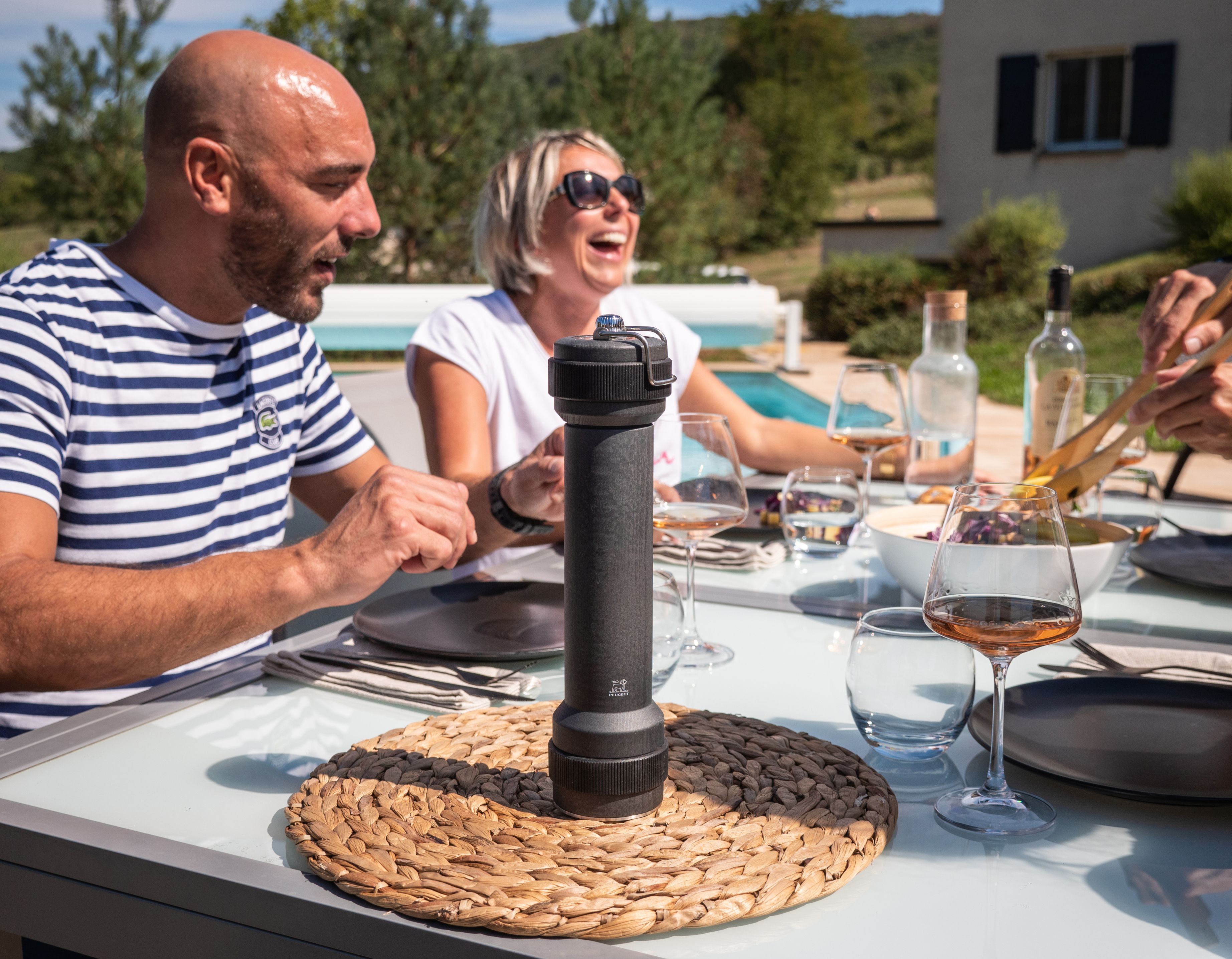 Peugeot BBQ Pfeffermühle in Graphit auf Esstisch mit lachenden Menschen im Hintergrund.