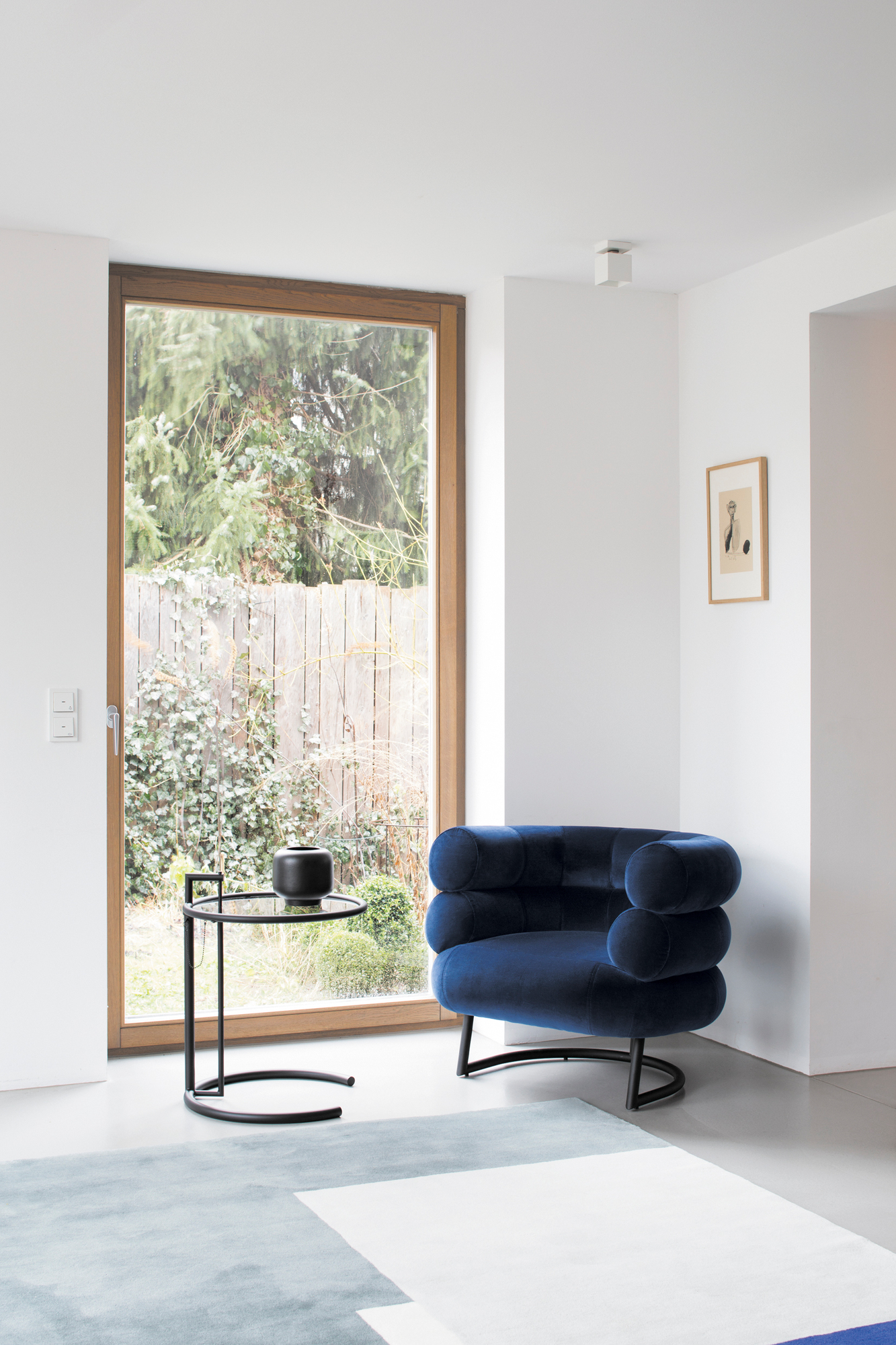 Adjustable Table E1027 Beistelltisch von ClassiCon neben einem blauen Sessel in einem hellen Wohnraum.