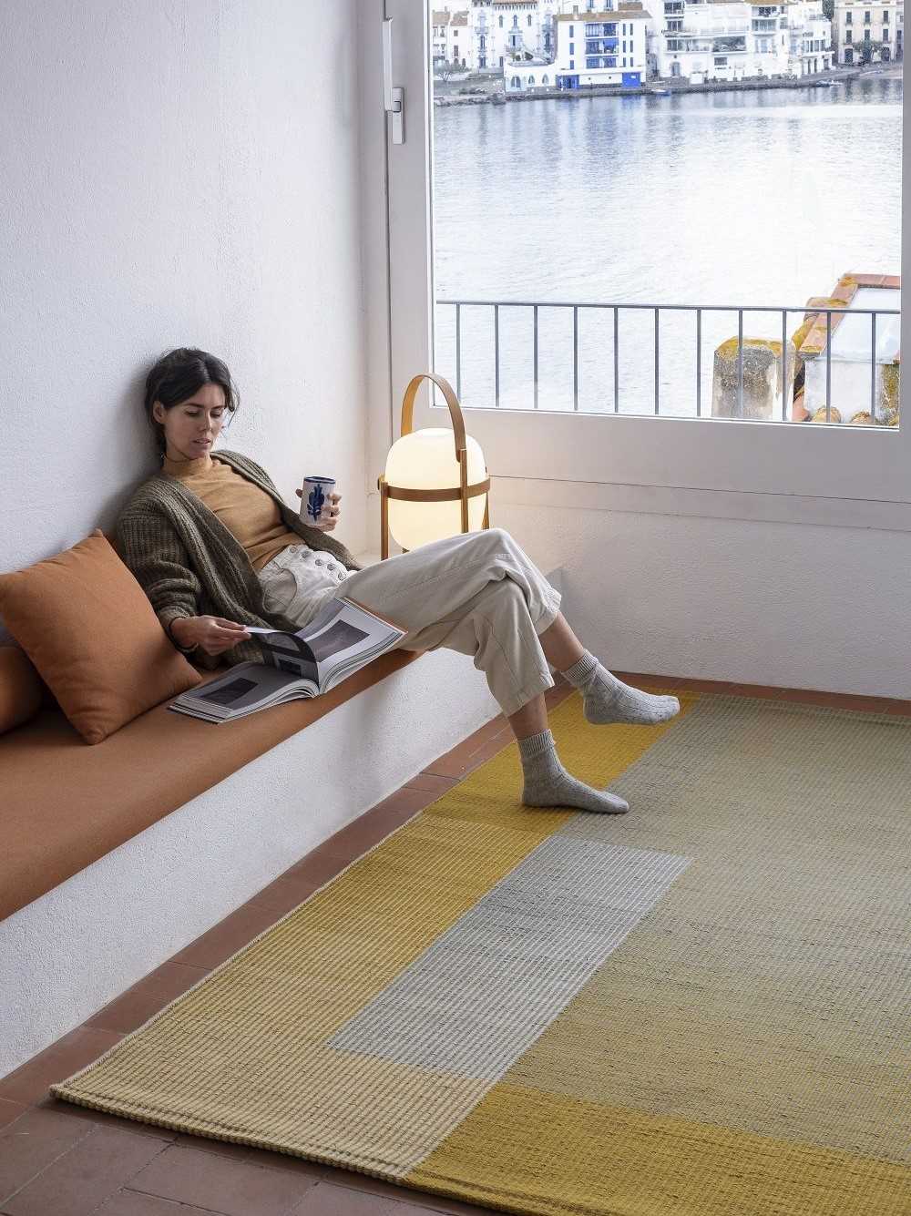 Frau liest auf einem gelb-grauen Teppich am Fenster mit Meerblick. Moderner Teppich für Wohnzimmer.