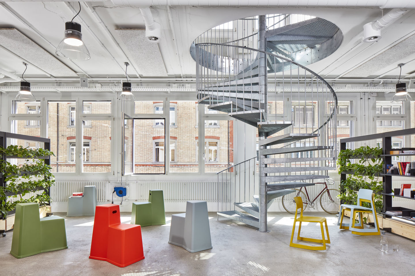 Büro mit bunten Tip Ton Stühlen von Vitra, spiralförmiger Treppe und großen Fenstern für moderne Arbeitsumgebung.