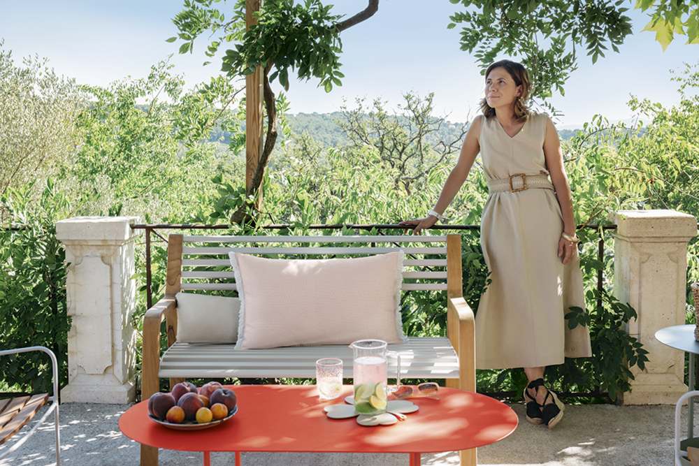 Frau lehnt an Somerset Bank von Fermob auf einer sonnigen Terrasse mit Gartenblick.