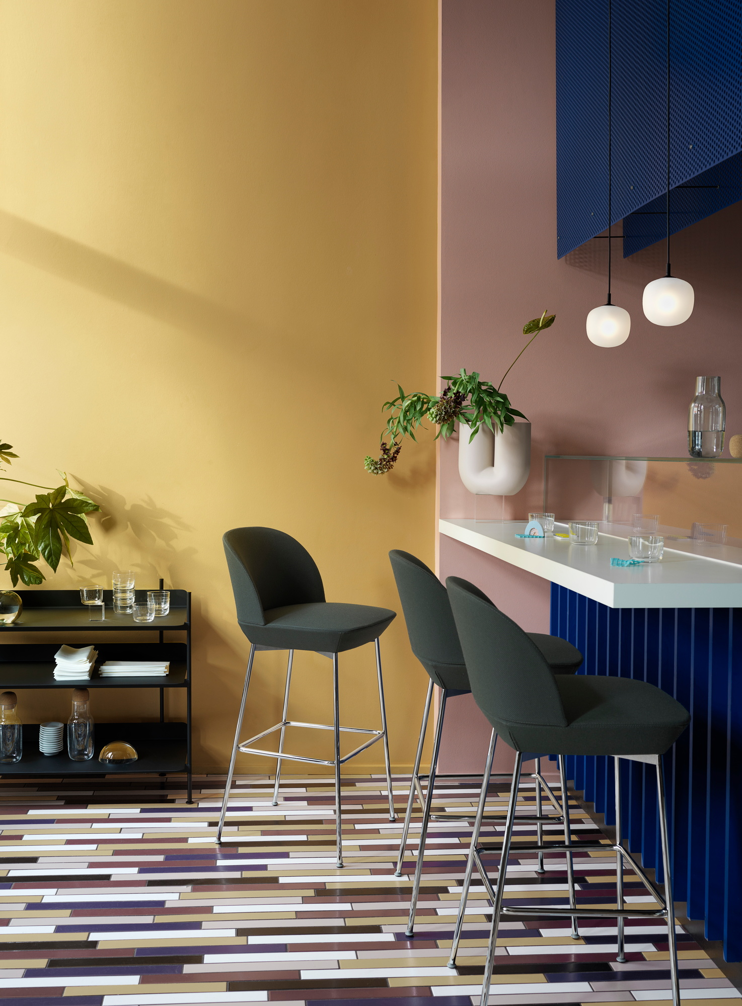 Drei Oslo Barhocker von Muuto in dunklem Grün mit Chromgestell vor farbenfrohem Hintergrund.