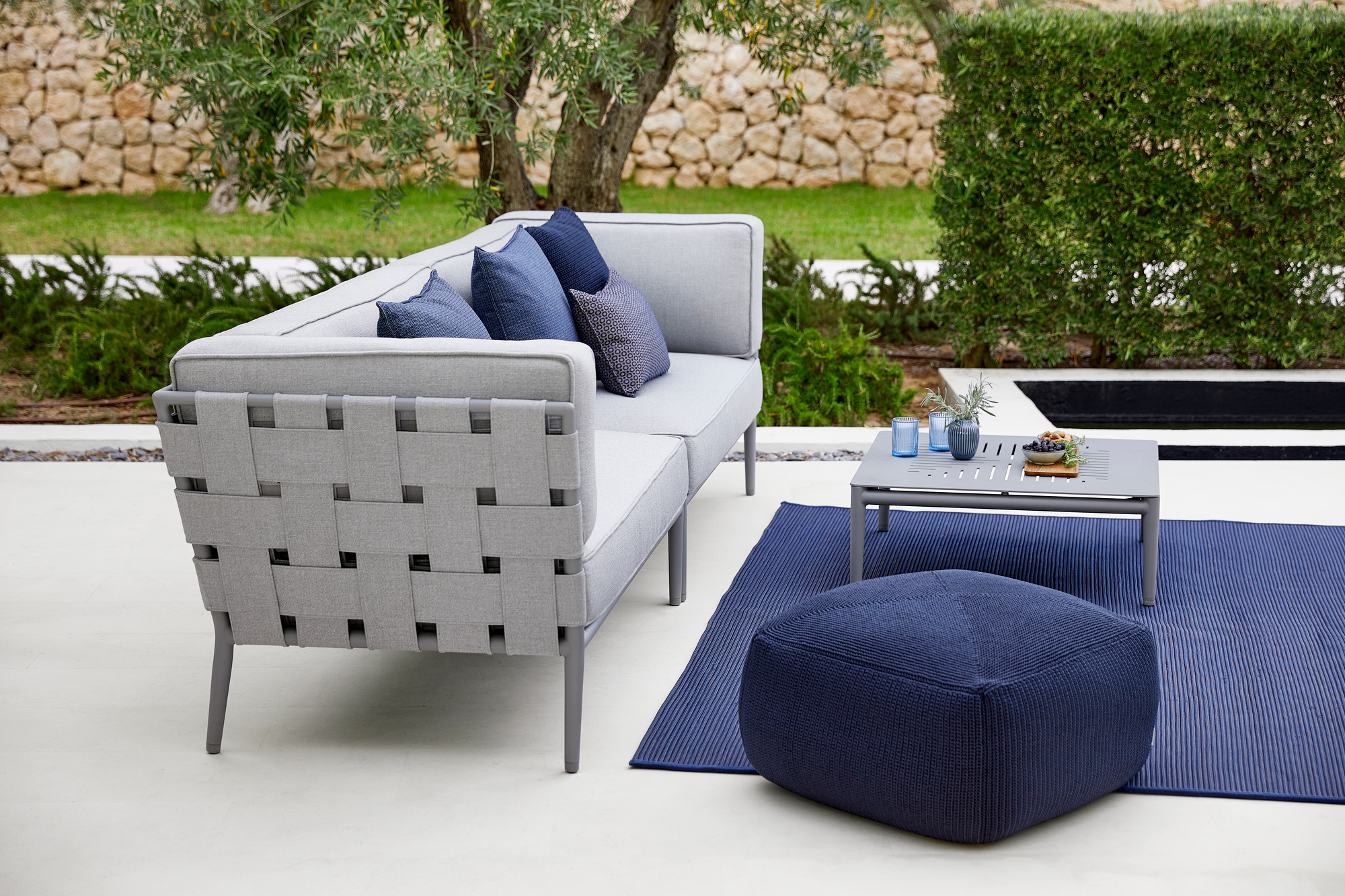 Hellgraues Conic AirTouch 2-Sitzer Outdoor Sofa mit blauen Kissen, Tisch und Hocker auf Terrasse.